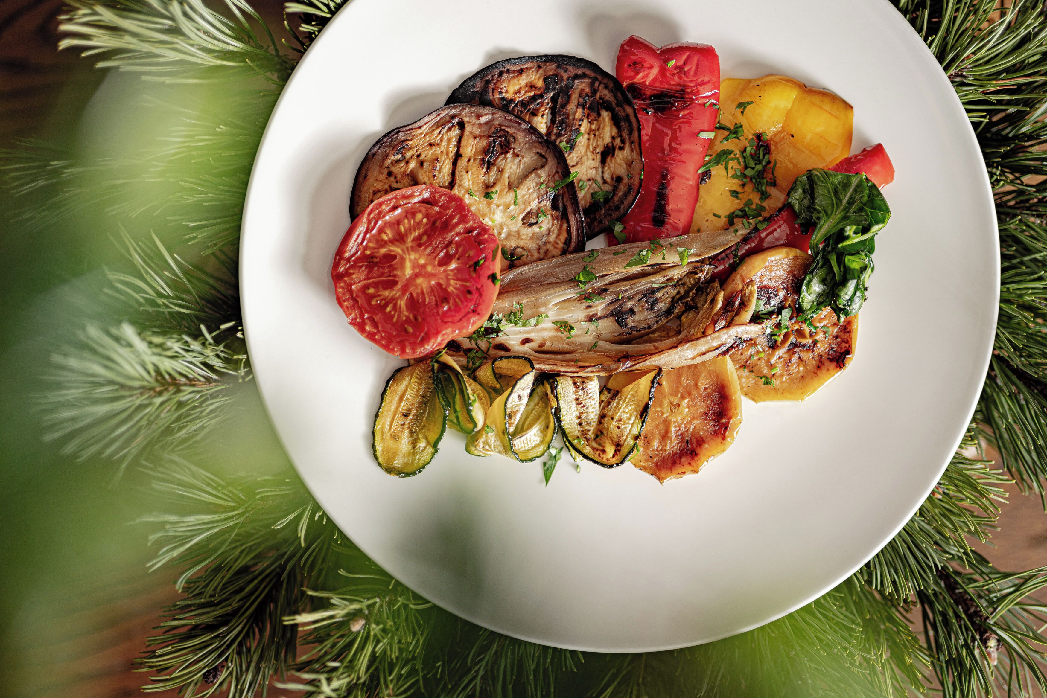 Ein Teller mit gegrilltem Gemüse, darunter Zucchini, Auberginen und Tomaten, garniert mit Kräutern, vor festlichem Hintergrund mit Tannenzweigen.