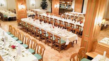 A banquet hall with multiple tables set for dining, wooden chairs, and a Christmas tree in the background.