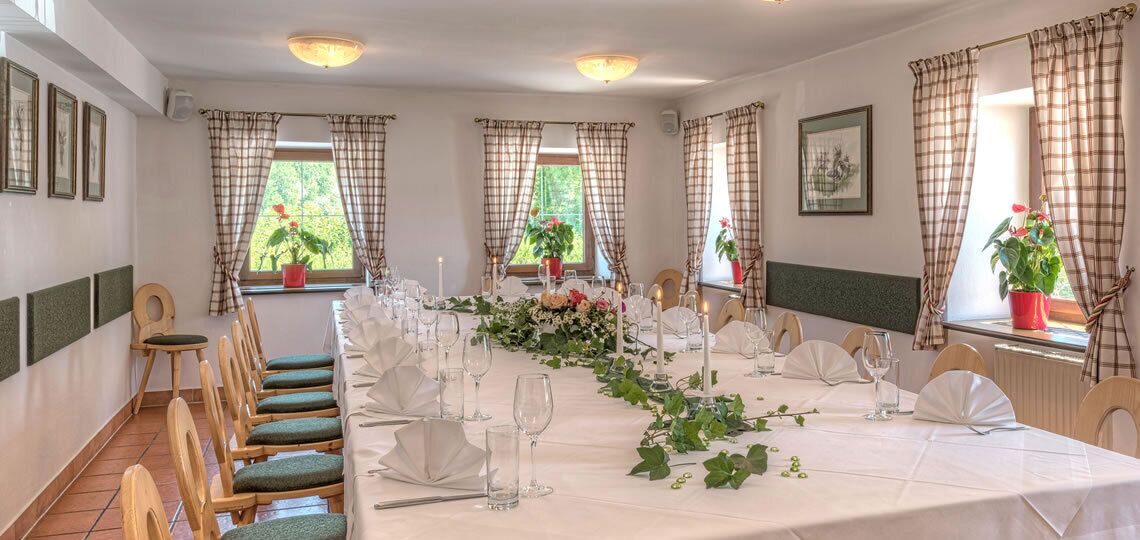 Ein Esszimmer mit einem langen Tisch, gedeckt für eine Mahlzeit, umrahmt von Stühlen, mit Fenstern und Vorhängen.