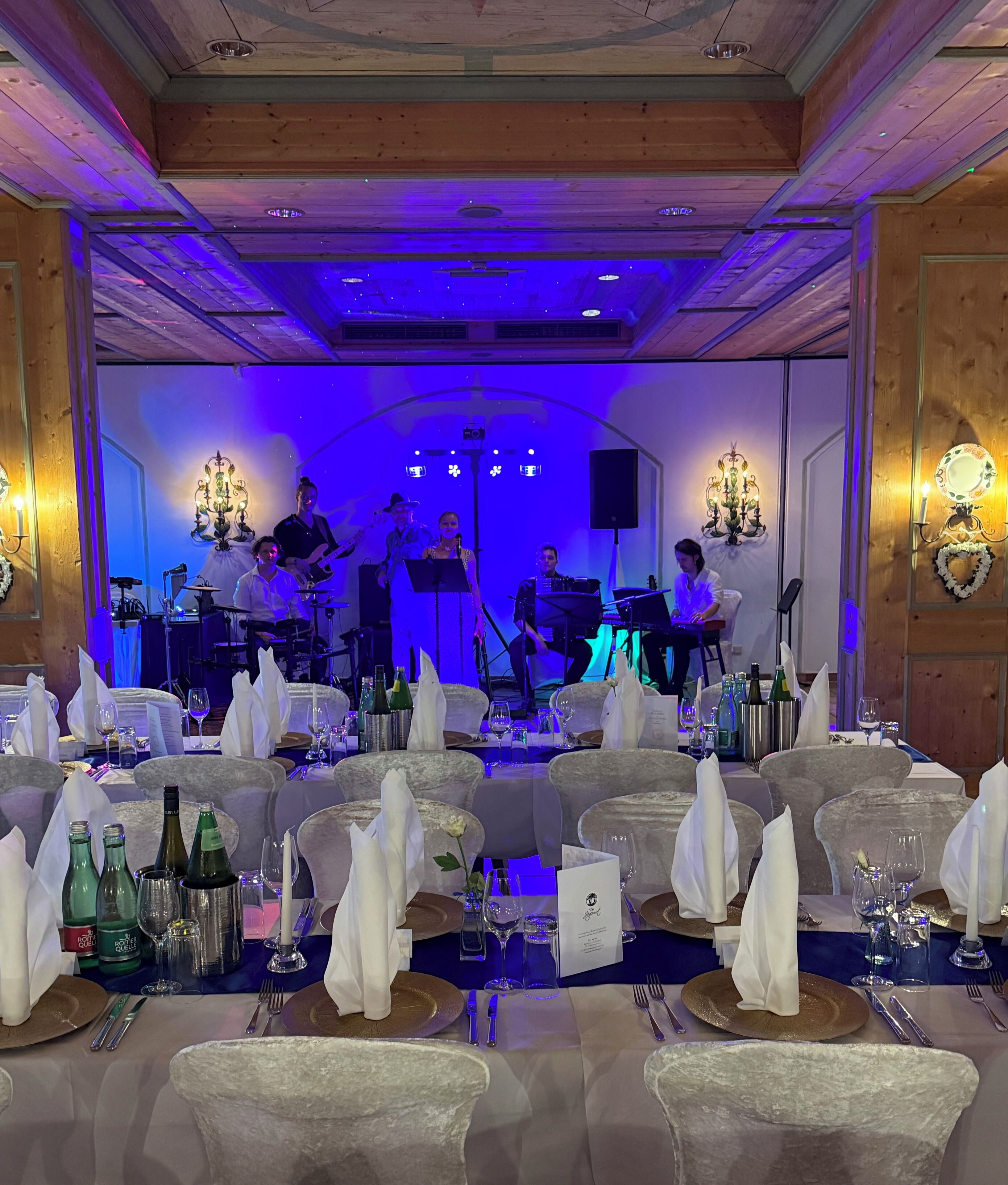 A formal dining area with tables set for a meal, a band performing on stage, and a wooden ceiling with blue lighting.