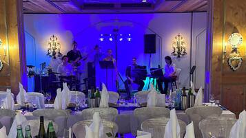 A formal dining area with tables set for a meal, a band performing on stage, and a wooden ceiling with blue lighting.