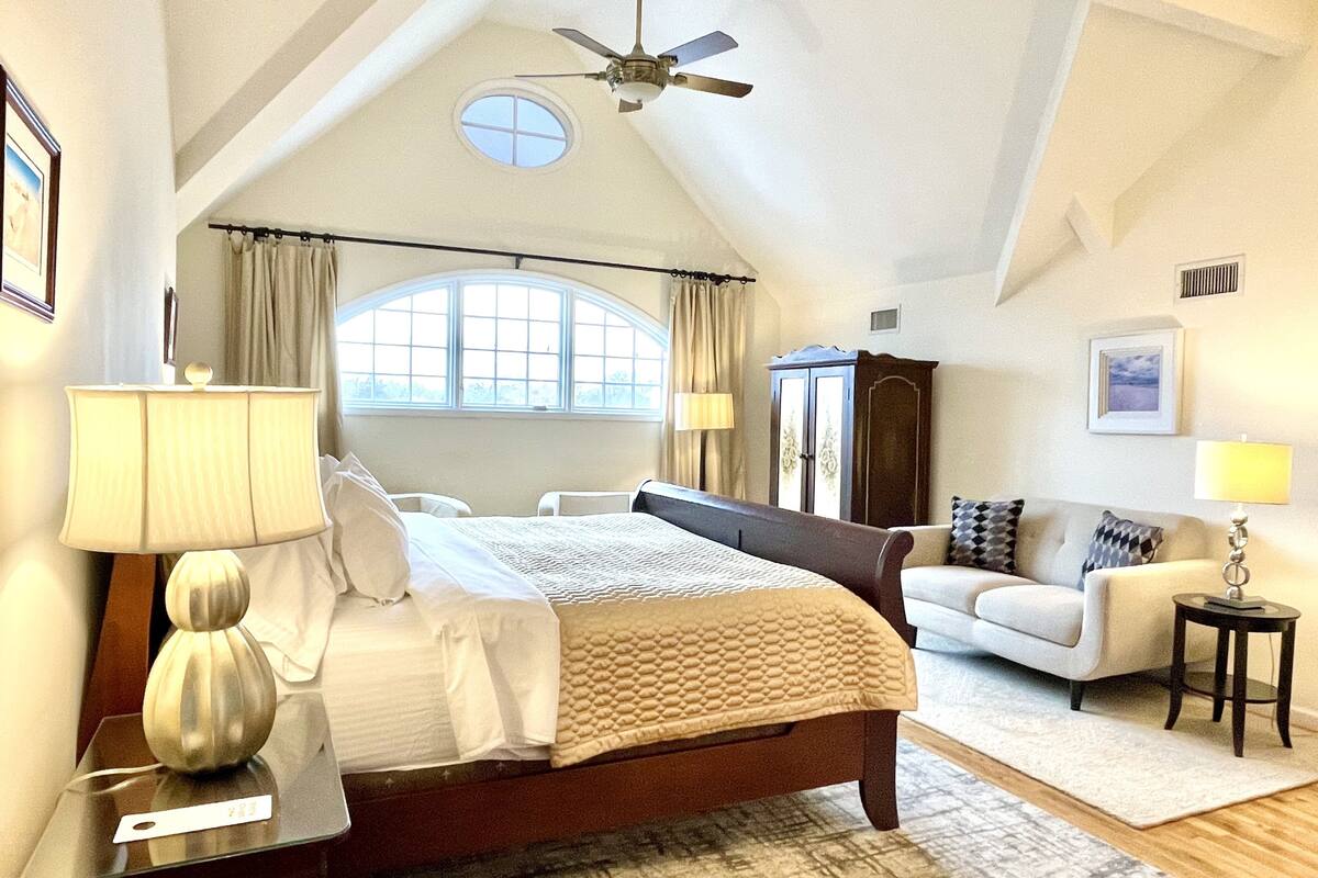 A bedroom with a bed, a nightstand, a lamp, a dresser, a window, a ceiling fan, and a view of the outdoors.