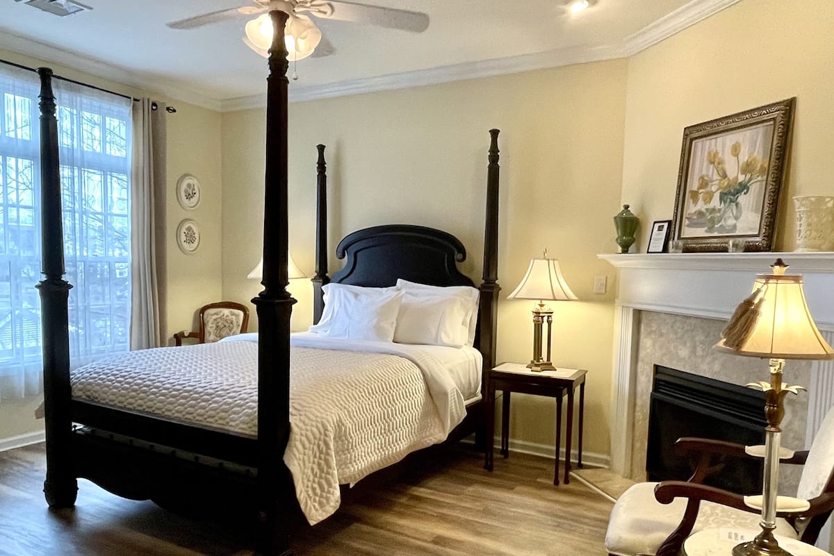A four-poster bed with a white bedspread, two bedside tables with lamps, a fireplace, and a large window with curtains.