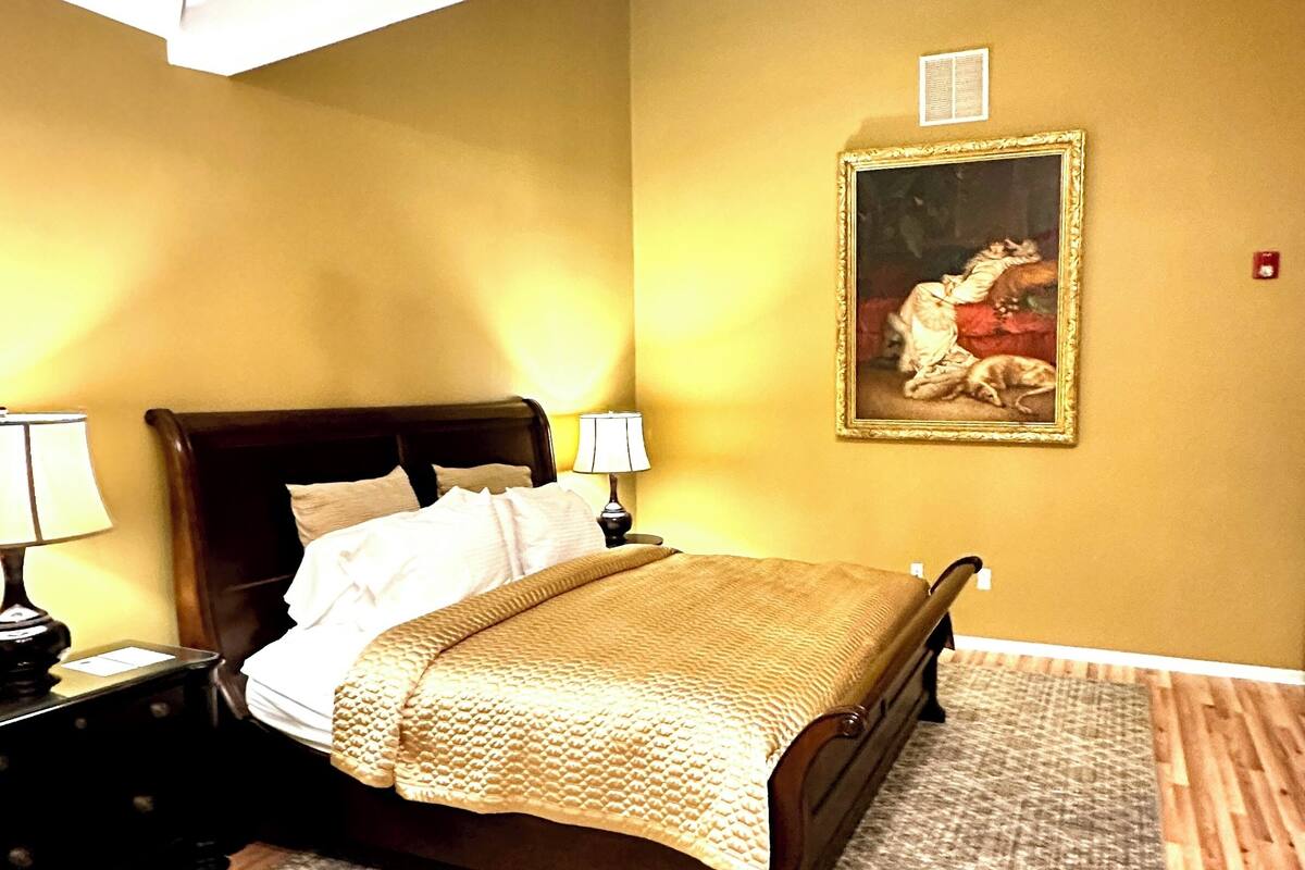 A hotel room with a bed, nightstands, a chandelier, a painting on the wall, and a patterned rug.