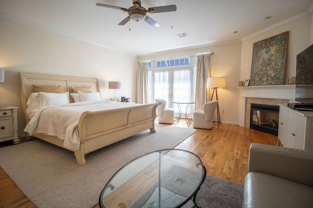 A bedroom with a bed, a glass table, a chair, a fireplace, and a ceiling fan.