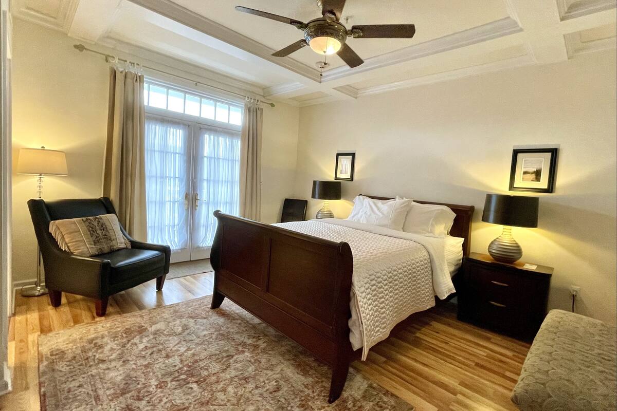A bedroom with a bed, a chair, a nightstand, a lamp, a ceiling fan, and a window with curtains.