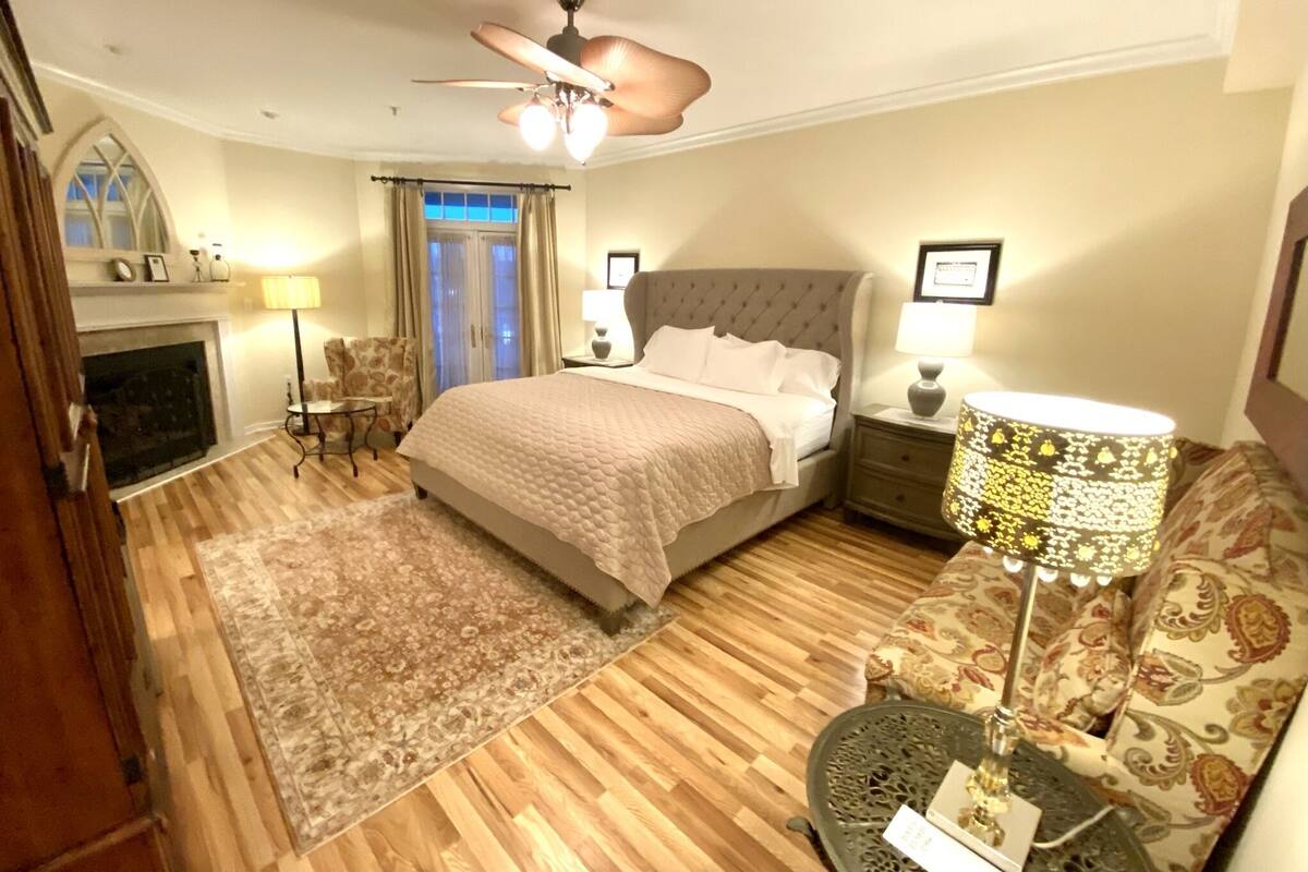 A bedroom with a bed, a chandelier, a fireplace, a sofa, and a patterned rug.