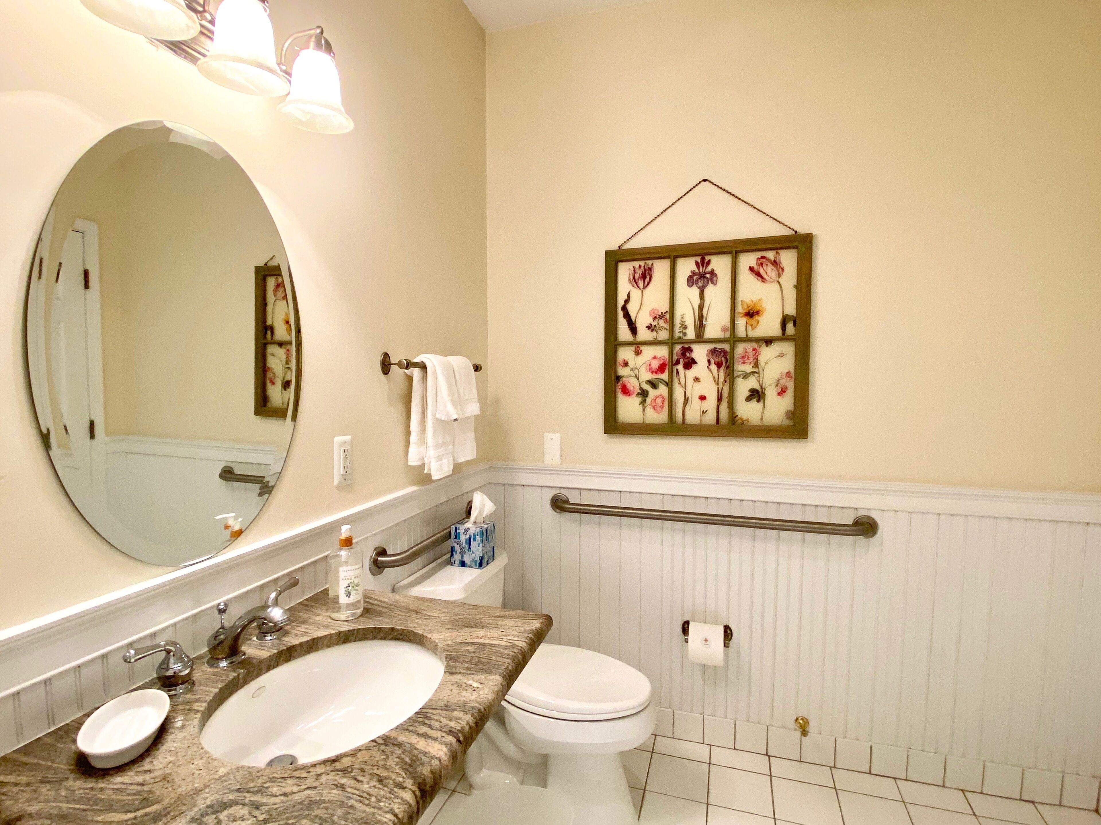 Un baño con lavamanos doble, inodoro, espejo y una obra de arte floral en el marco en la pared.