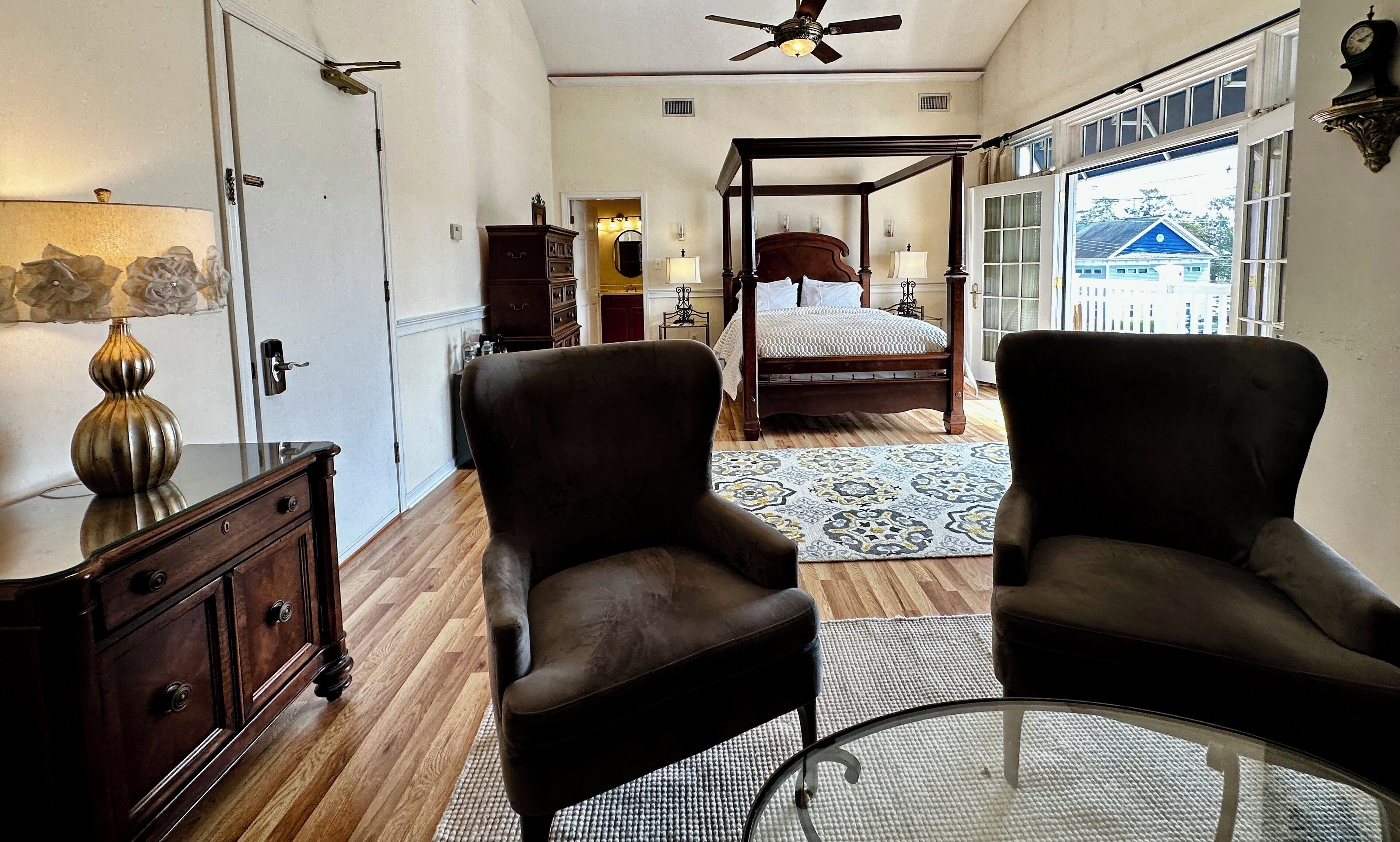 A bedroom with a four-poster bed, a sitting area with two armchairs, a wooden dresser, and a lamp.