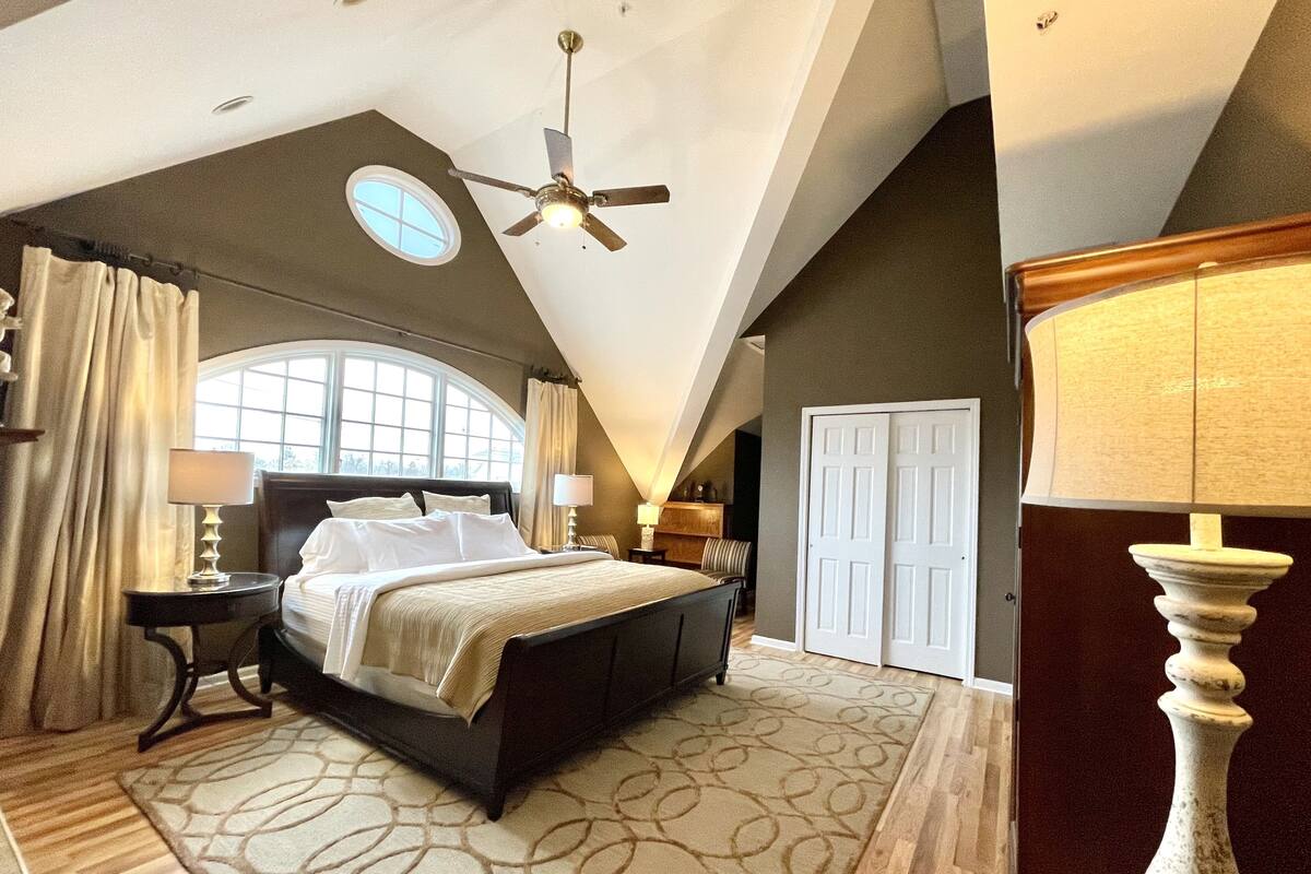 A bedroom with a large bed, a ceiling fan, a window, and a door.