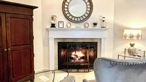Un salon avec une cheminée, deux fauteuils, un meuble en bois et un miroir au-dessus du manteau de cheminée.