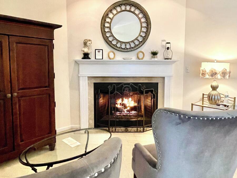 A living room with a fireplace, two armchairs, a wooden cabinet, and a mirror above the mantel.