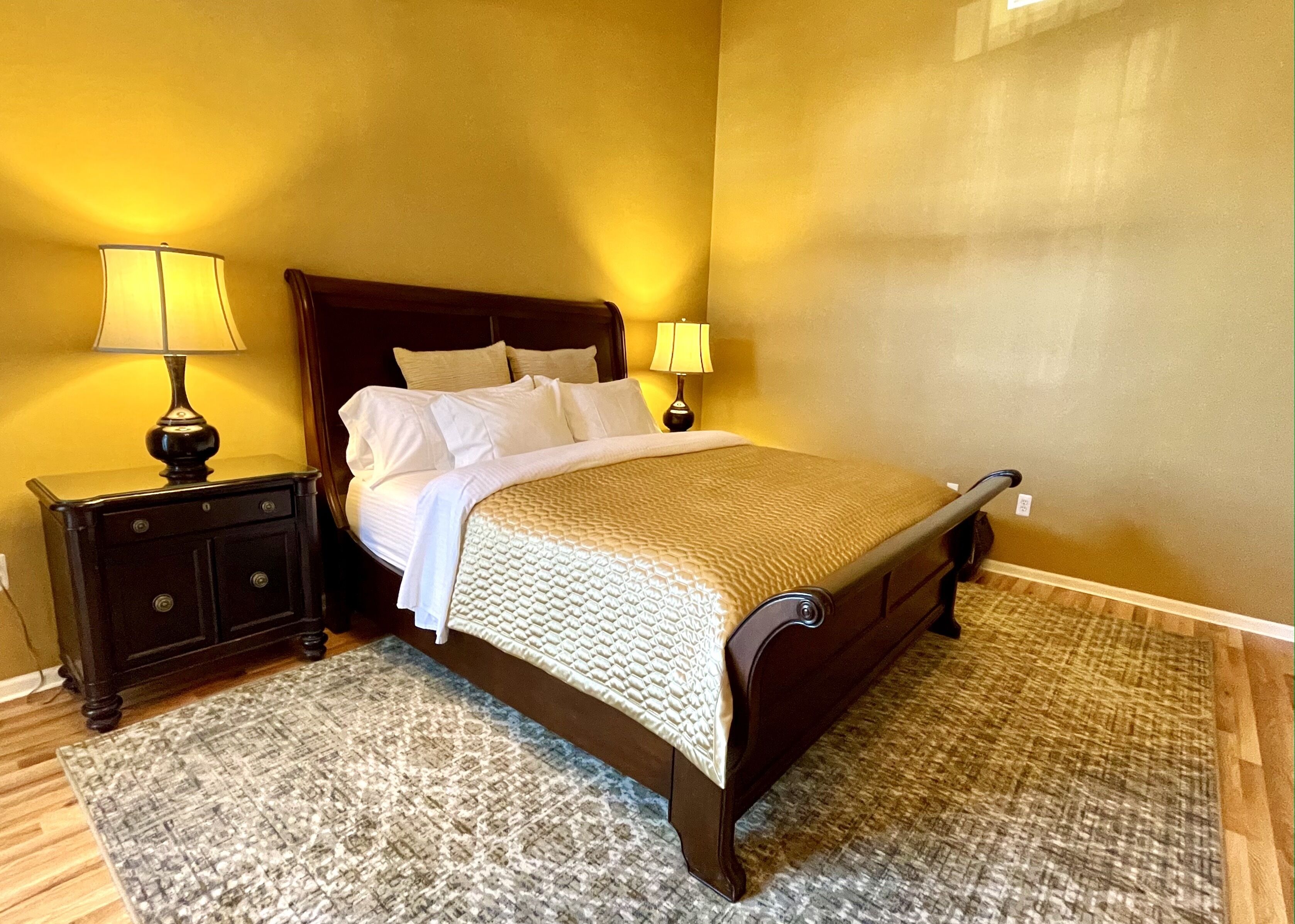 A neatly made bed with a wooden headboard, two bedside tables with lamps, and a patterned rug on a wooden floor.