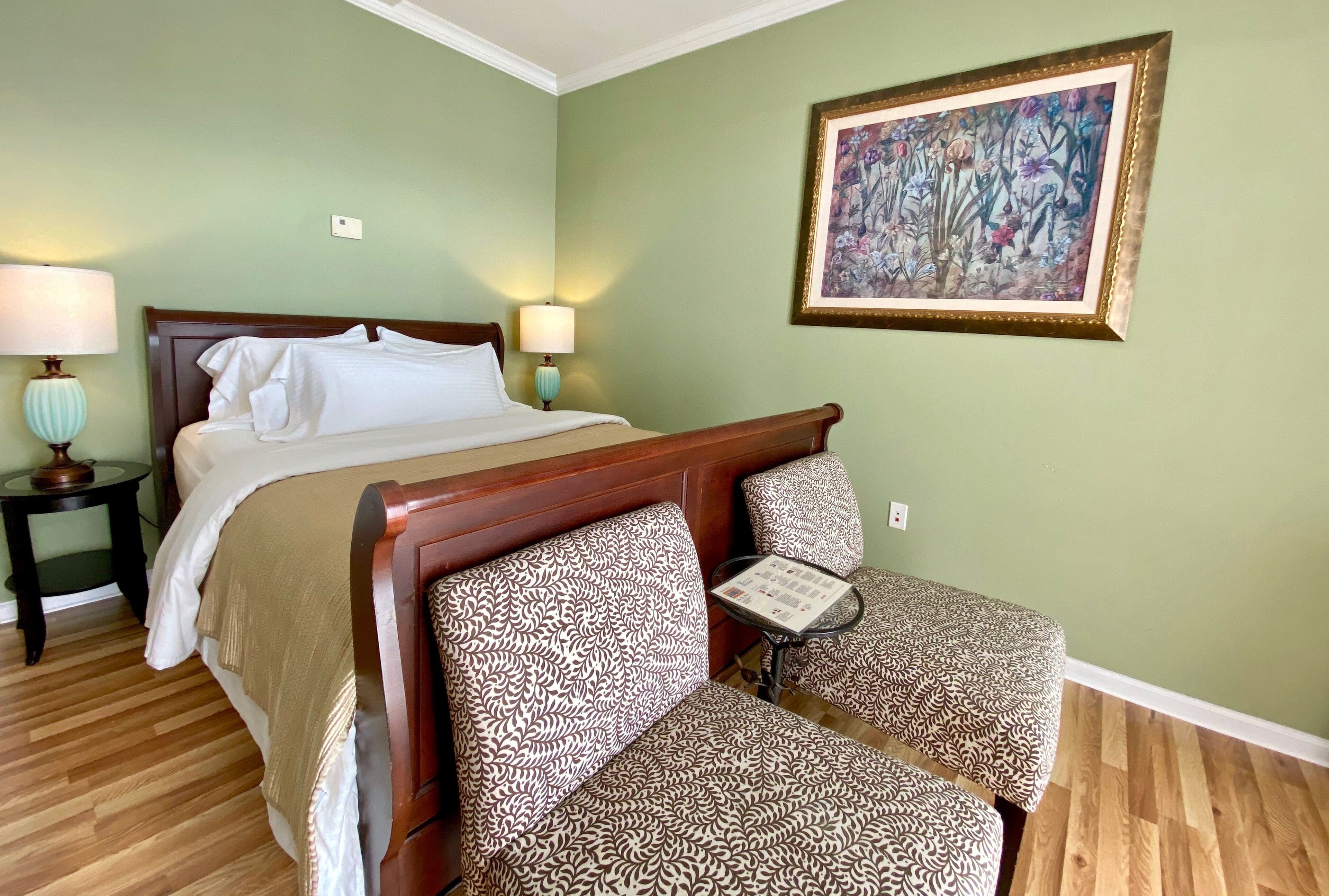 Ein ordentlich eingerichtetes Hotelzimmer mit einem Bett, zwei gemusterten Stühlen, einem kleinen Tisch und einer gerahmten Blumenleiste an der Wand.