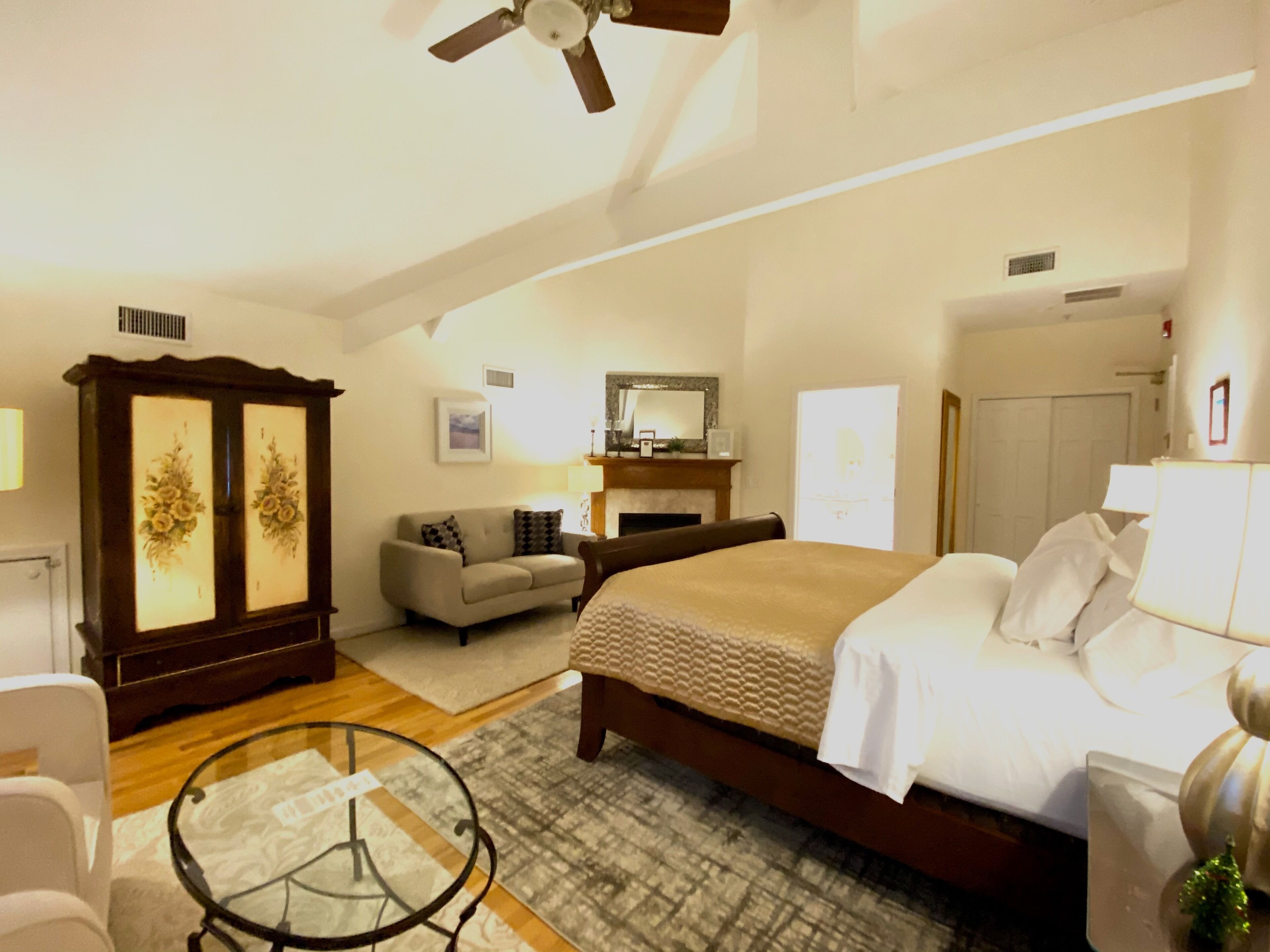 Een ruime slaapkamer met een groot bed, een houten garderobe, een bank, een glazen salontafel en een plafondventilator.