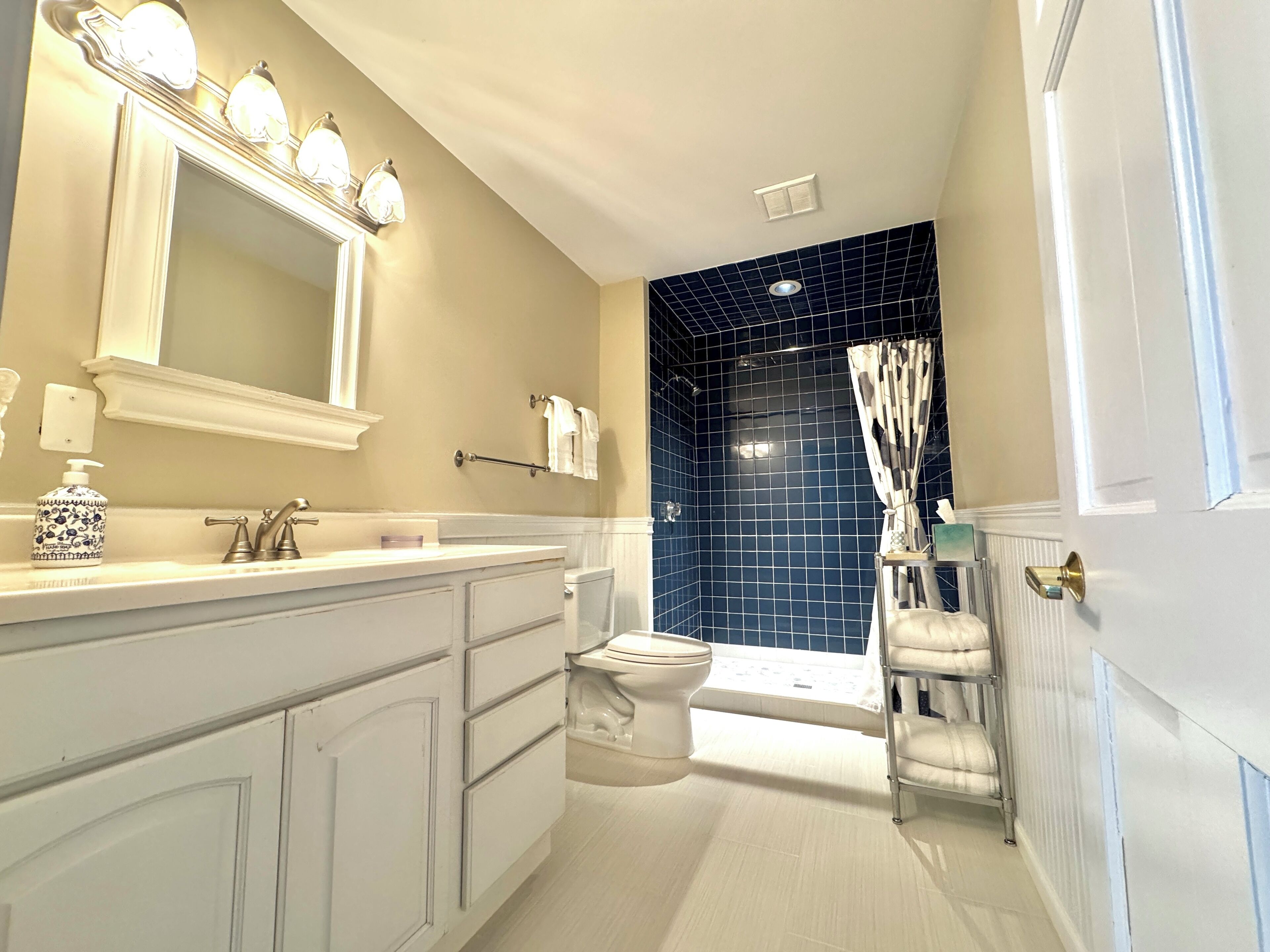 A bathroom with a white vanity, a large mirror with lights, a blue-tiled shower area, and a white toilet.