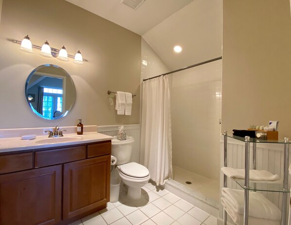 A bathroom with a wooden vanity, a round mirror, a toilet, a glass shower enclosure, and a glass shelf with towels.