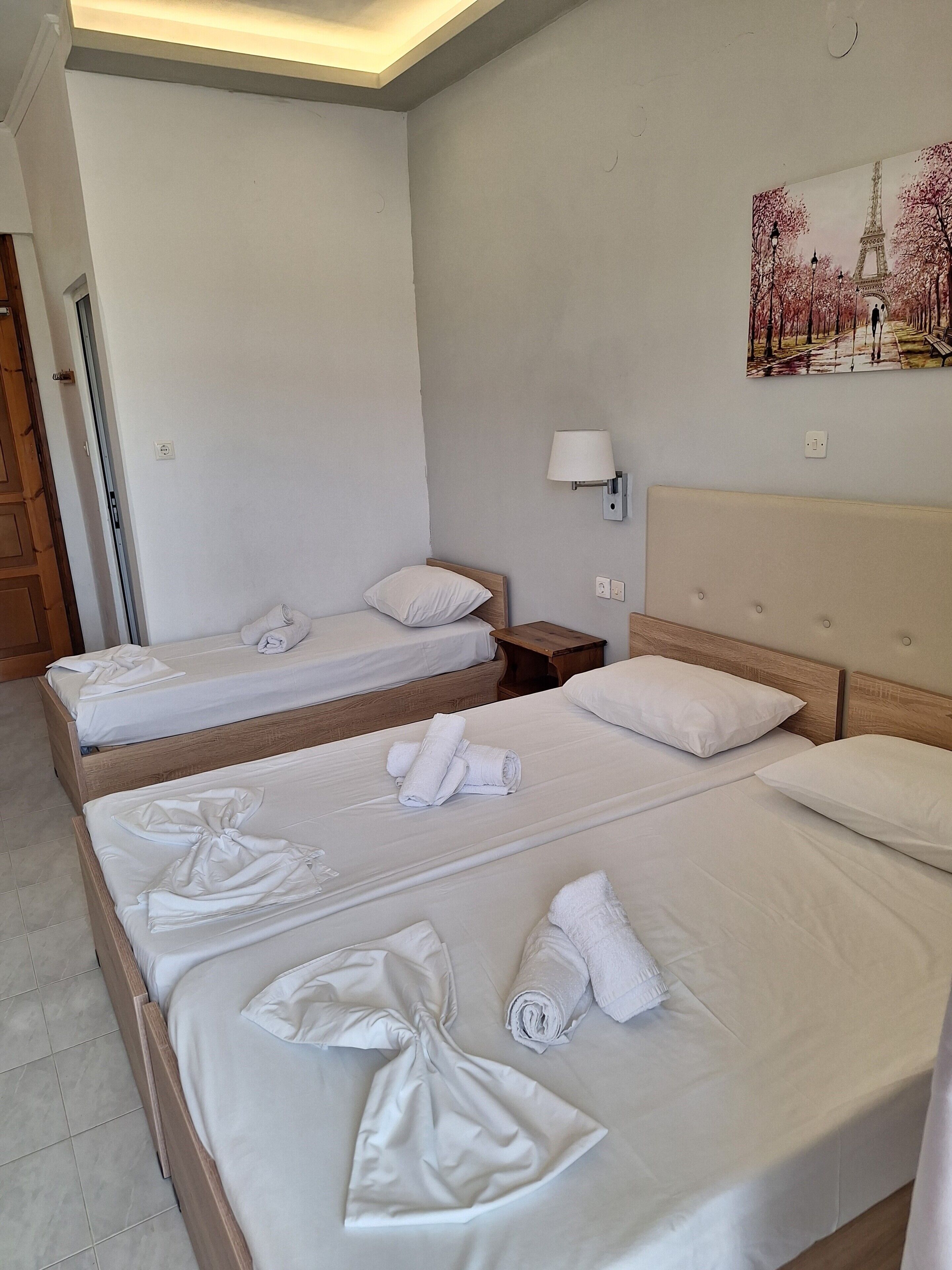 Quarto de hotel com duas camas, uma mesa de cabeceira, uma luminária na parede e um quadro da Torre Eiffel.