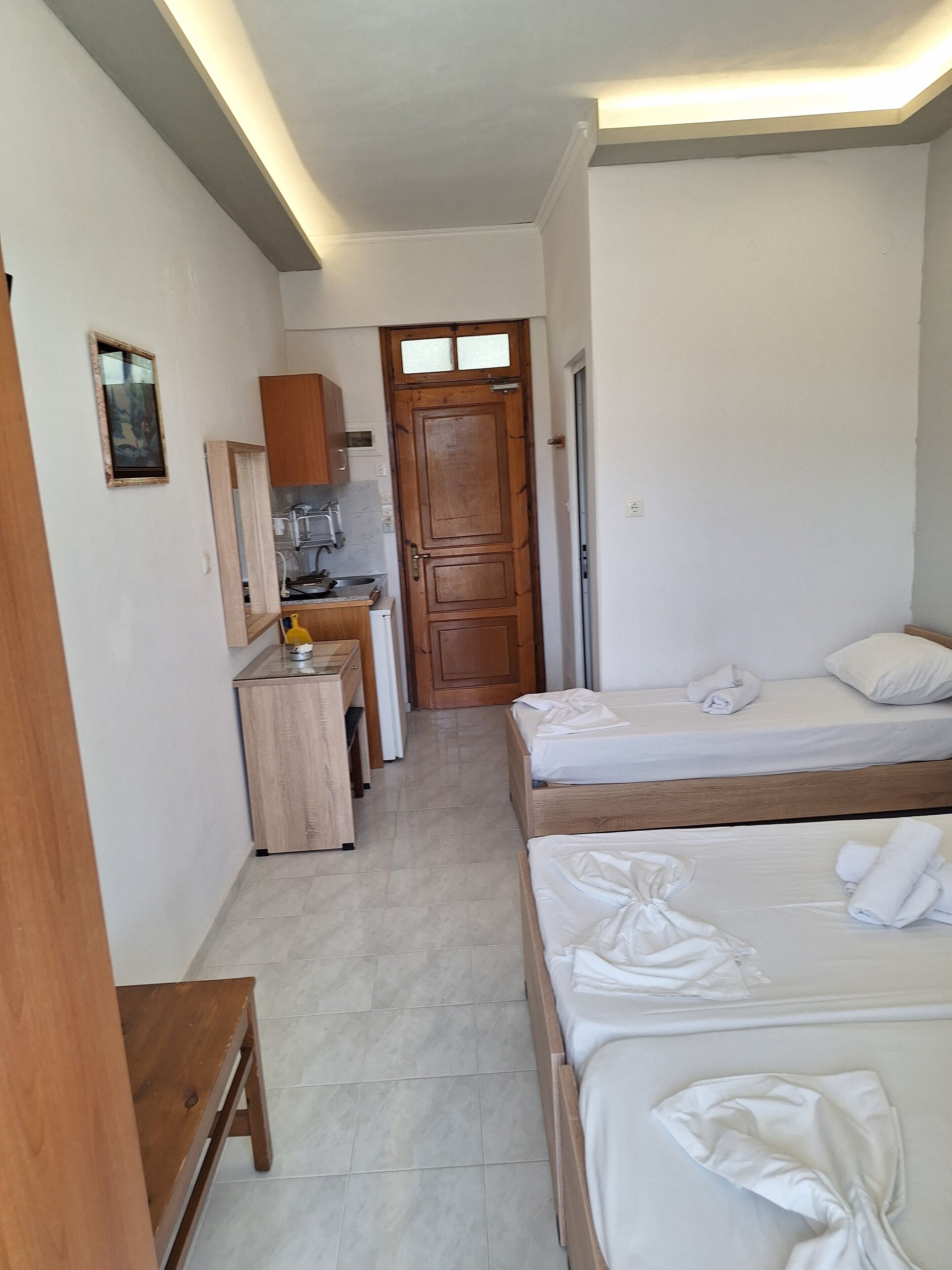 A narrow room with two beds, a small kitchenette, and a wooden door.