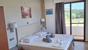 A hotel room with a bed, a nightstand, a lamp, a wardrobe, a window with a view, and a wall with two pictures. - Vera Studios (Rhodes)