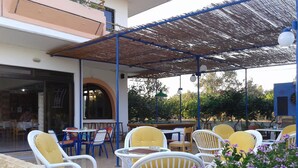 An outdoor patio with yellow and white wicker chairs, a covered area with a blue metal frame, and a view of the street and greenery in the background. - Vera Studios (Rhodes)