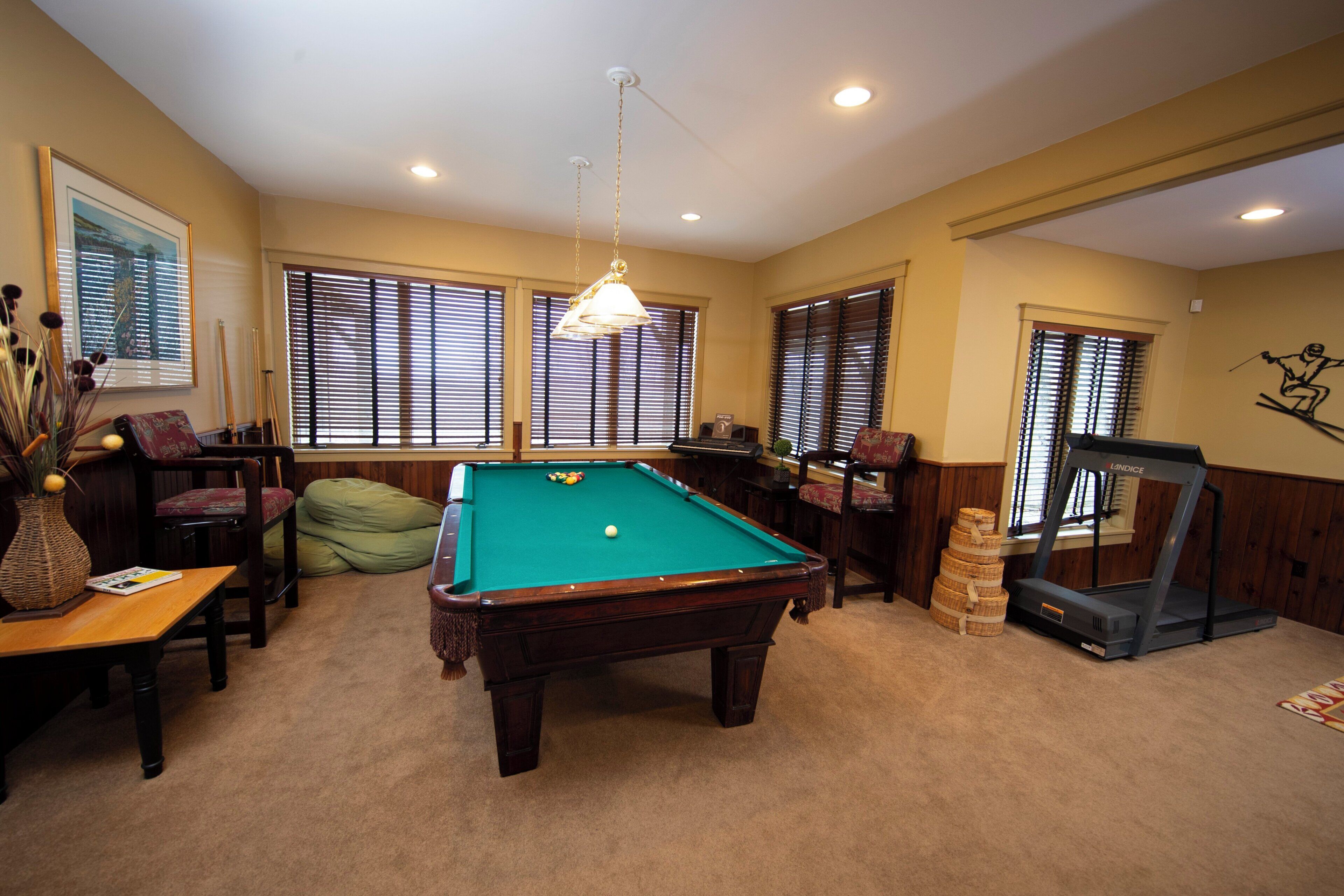 A pool table in a room with large windows, a treadmill, and a chair.