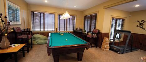 A pool table in a room with large windows, a treadmill, and a chair.