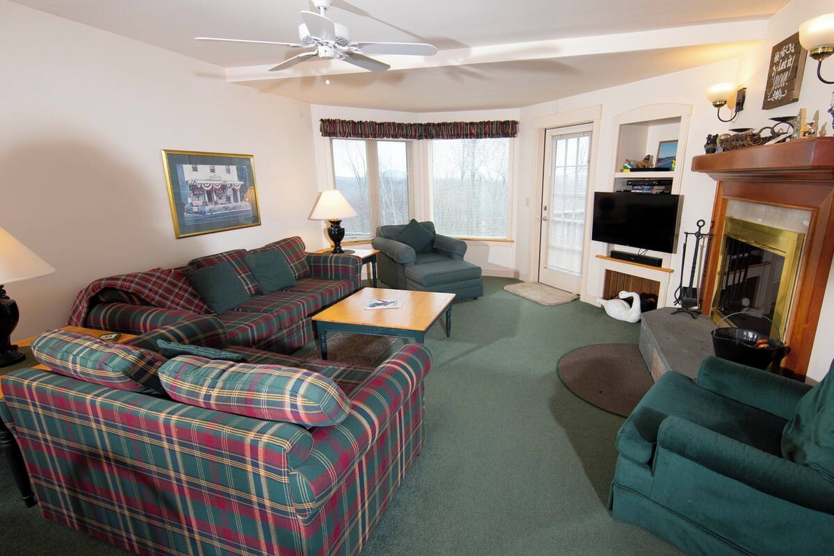 A cozy living room with a plaid sofa, a green armchair, a wooden coffee table, and a fireplace.
