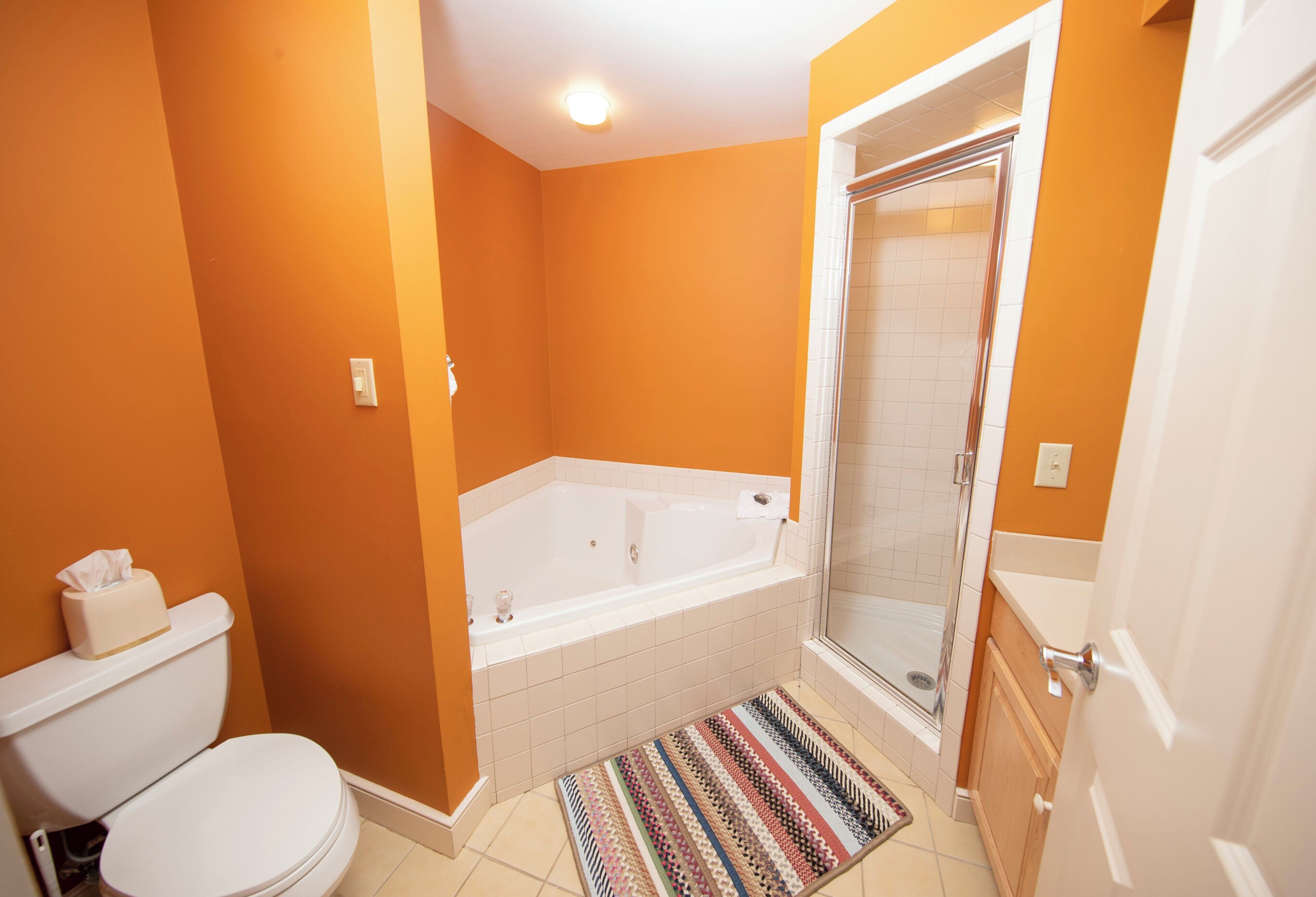 Un baño con una pared naranja, una bañera blanca, un cerramiento de ducha de vidrio y una alfombra estampada.