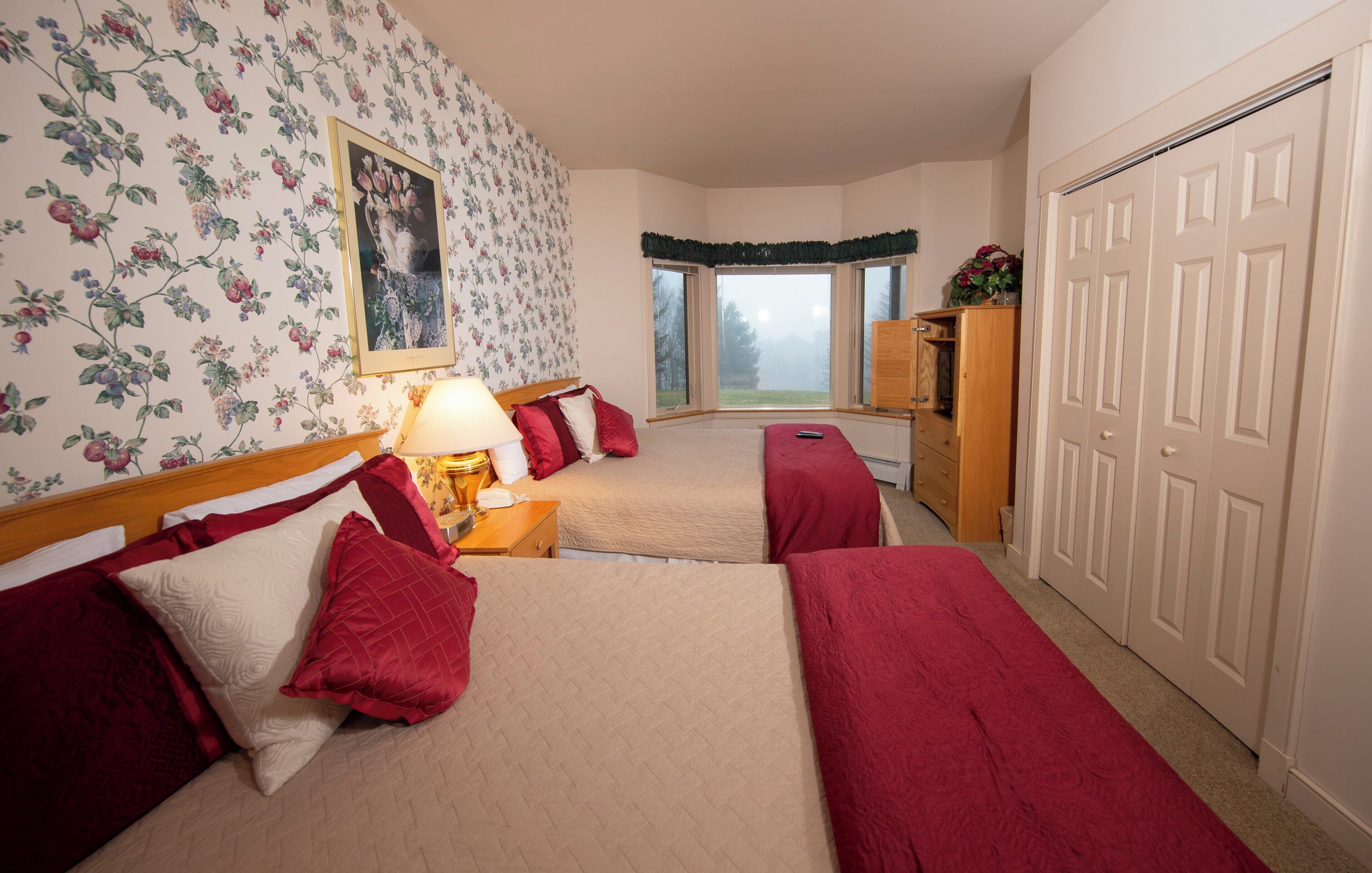 A bedroom with two beds, floral wallpaper, a window with a view, a wardrobe, and a desk with a lamp.