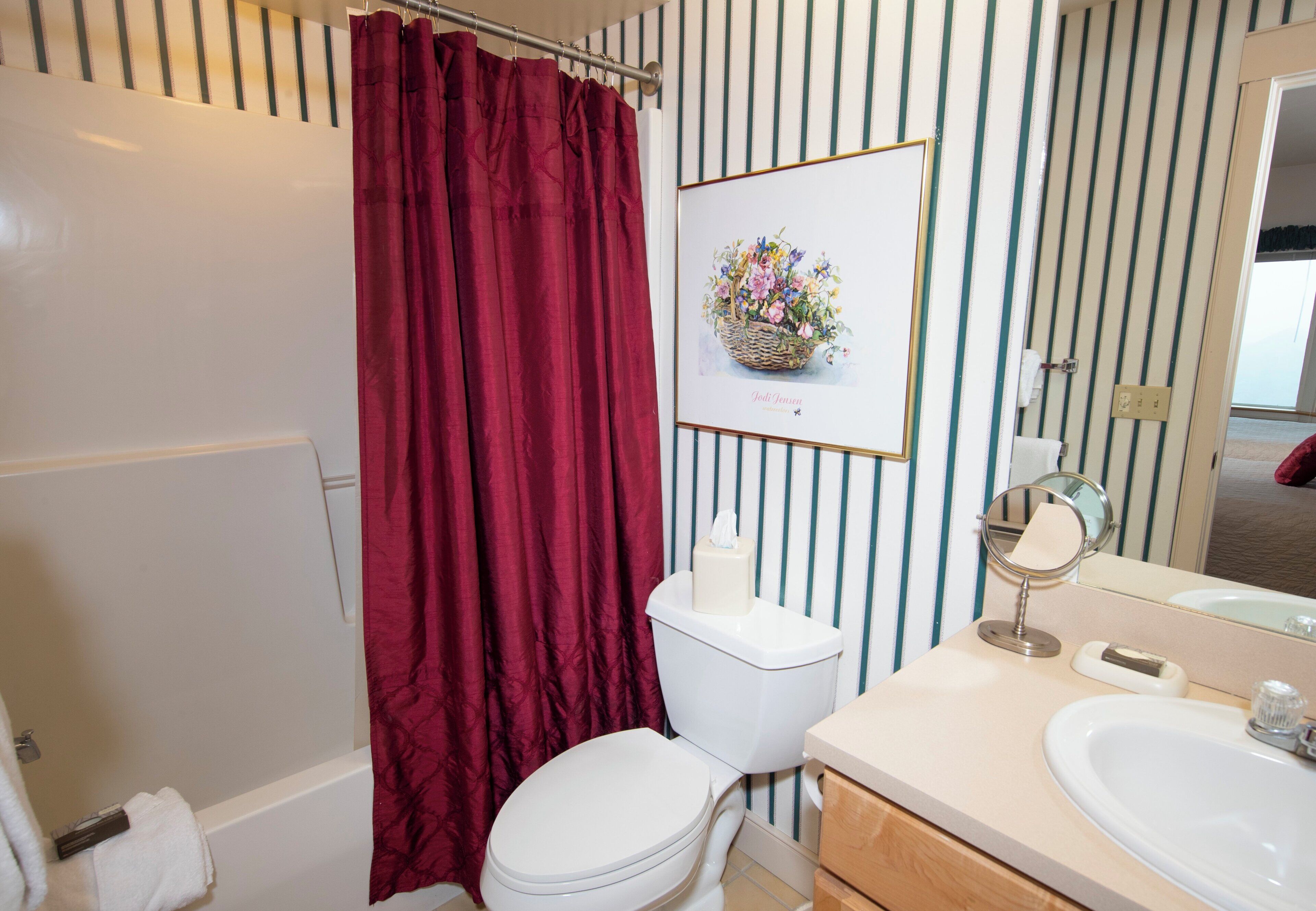 Un bagno con un water bianco, una tenda da doccia rossa, un quadro floreale appeso al muro, uno specchio piccolo e un lavandino con piano d'appoggio beige.