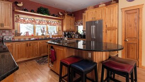 A kitchen with wooden cabinets, a stainless steel refrigerator, a large island with stools, and a window with decorative curtains. - Solitude Village at Okemo  by Vail Resorts (Ludlow)
