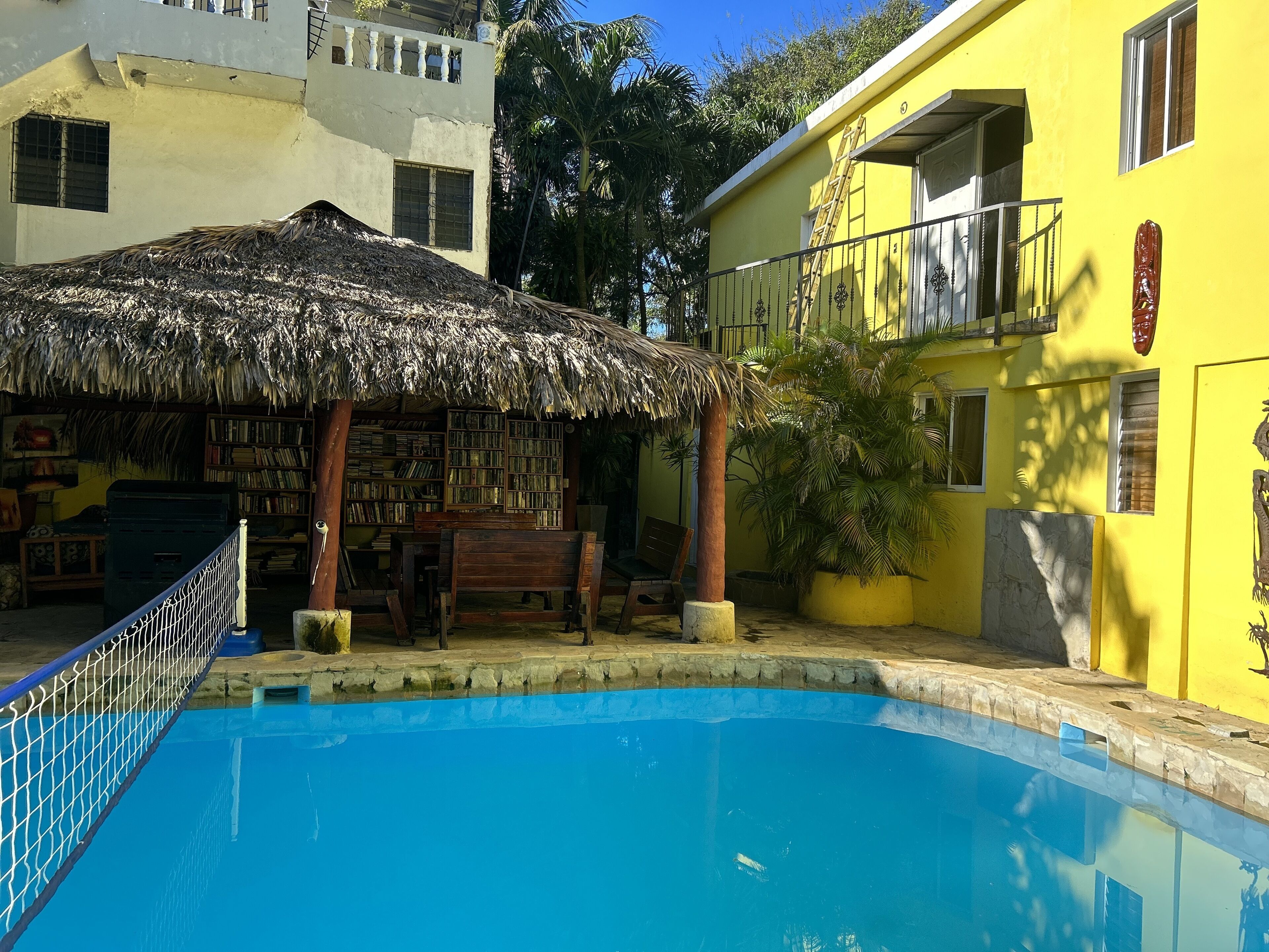 A pool area with a thatched-roof structure, bookshelves, and a yellow building with a balcony.