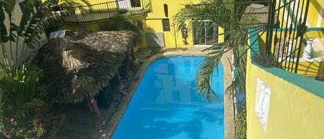 A yellow building with a swimming pool, a thatched-roof structure, and a fenced area with plants.