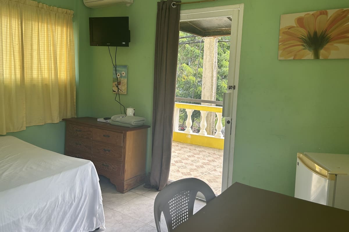 Una habitación con una cama, un aparador de madera, una mesa, un televisor montado en la pared y una puerta que da a un espacio exterior con barandilla amarilla.
