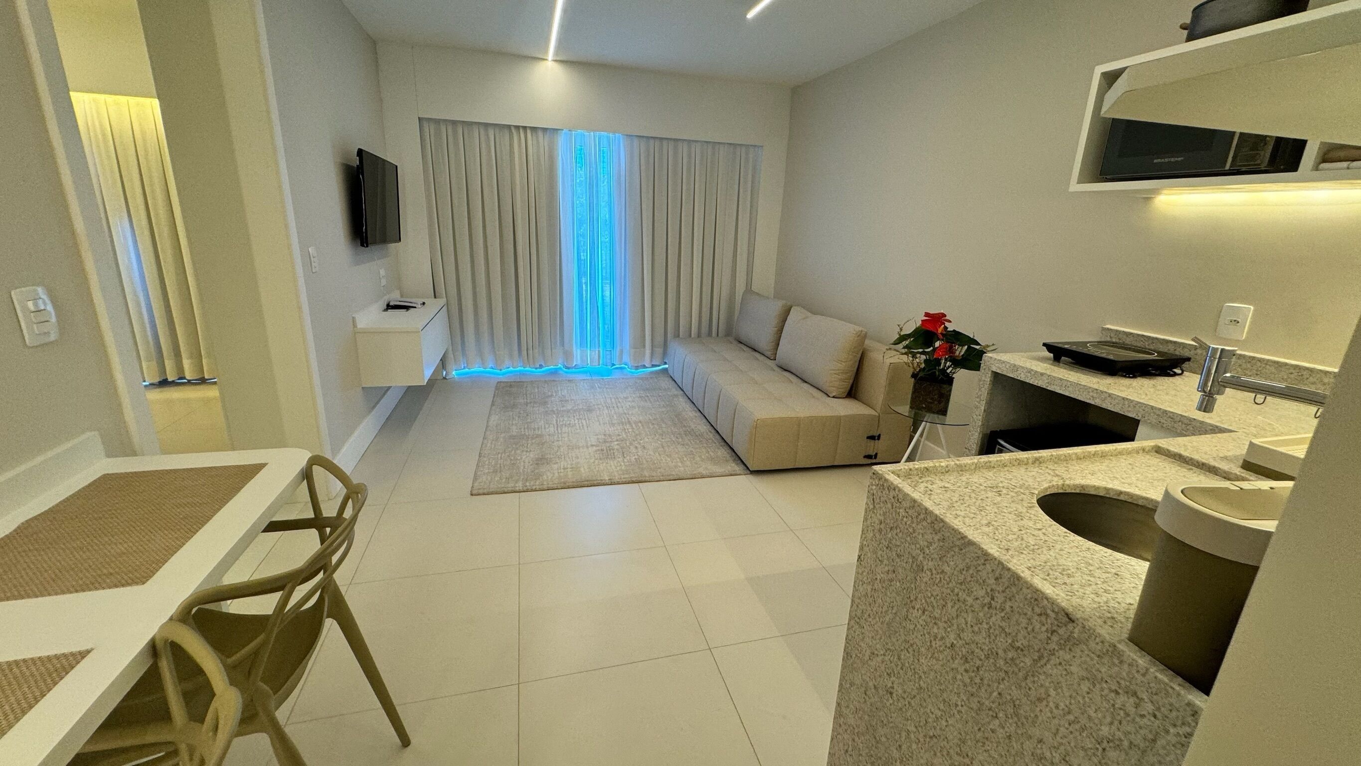 A modern living room with a sofa, a dining table with chairs, a kitchen area with a sink and countertop, and a TV mounted on the wall.