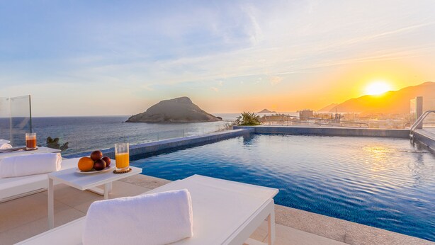 A poolside area with lounge chairs, a table with fruit and drinks, and a view of the ocean and sunset. - CDesign Hotel (Rio de Janeiro)