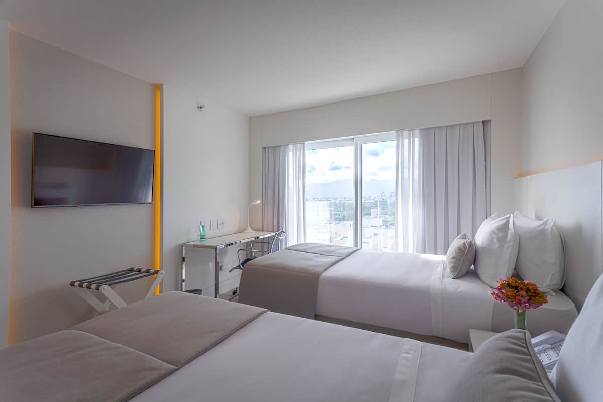 Habitación de hotel con dos camas, un escritorio, una silla, un televisor y una ventana con vista a la ciudad.