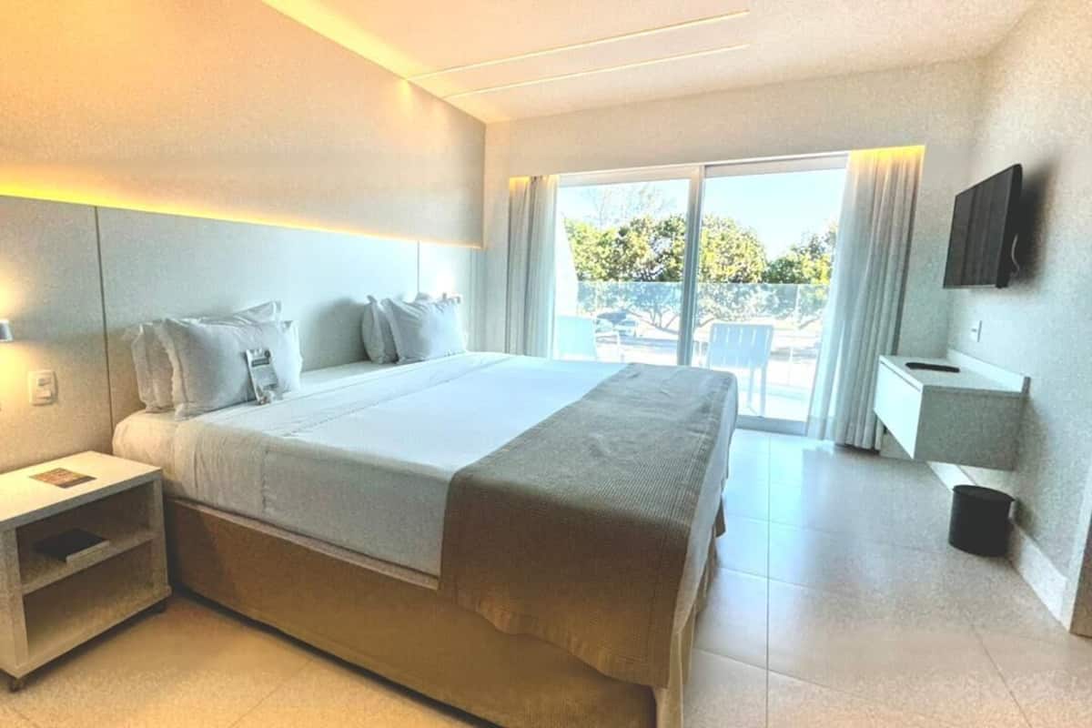 Una habitación de hotel moderna con una cama grande, mesitas de noche, televisión y un balcón con vistas.