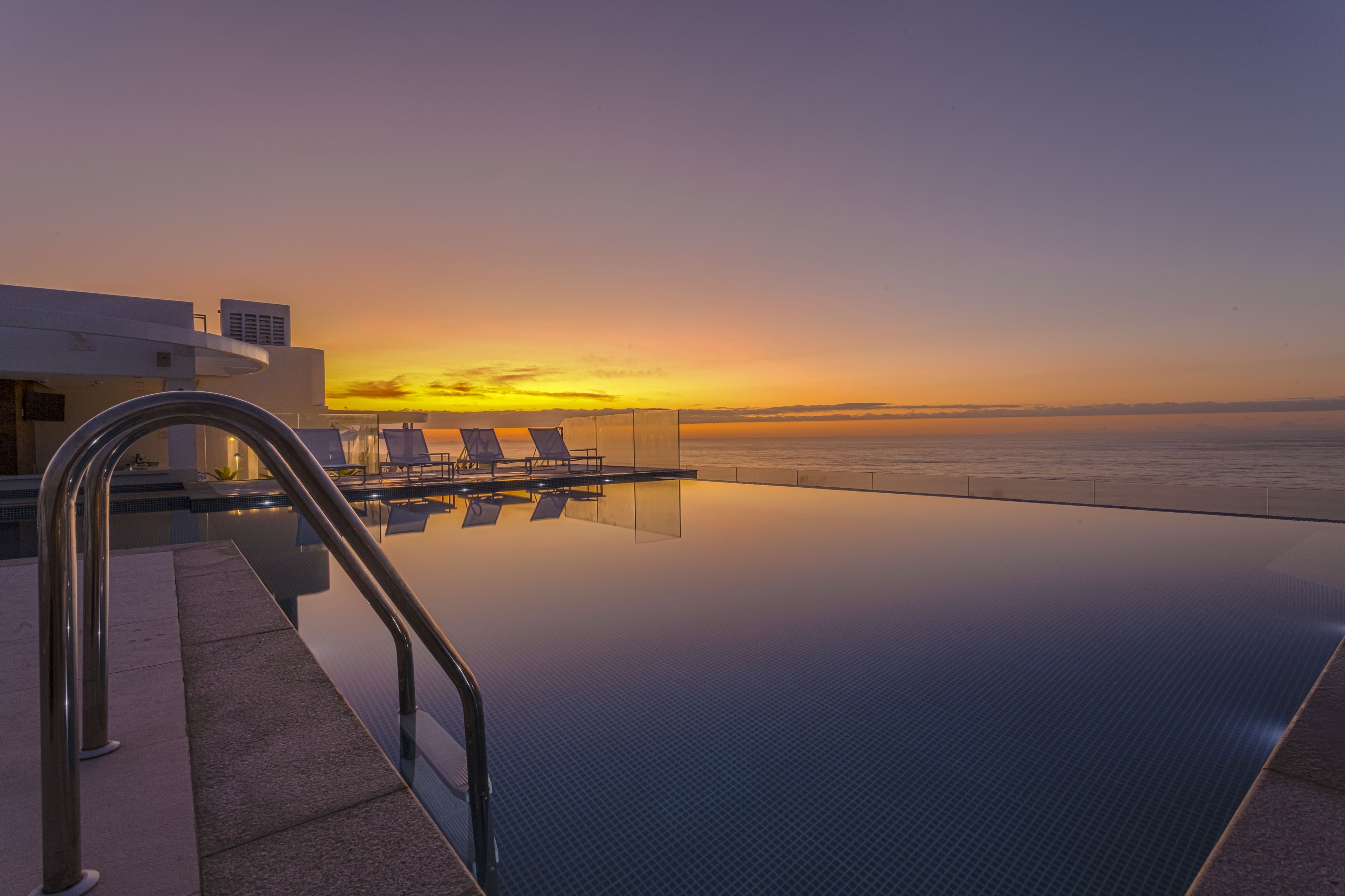 Una piscina infinita con vista al atardecer sobre el océano, sillones de descanso y una estructura de edificio moderna.