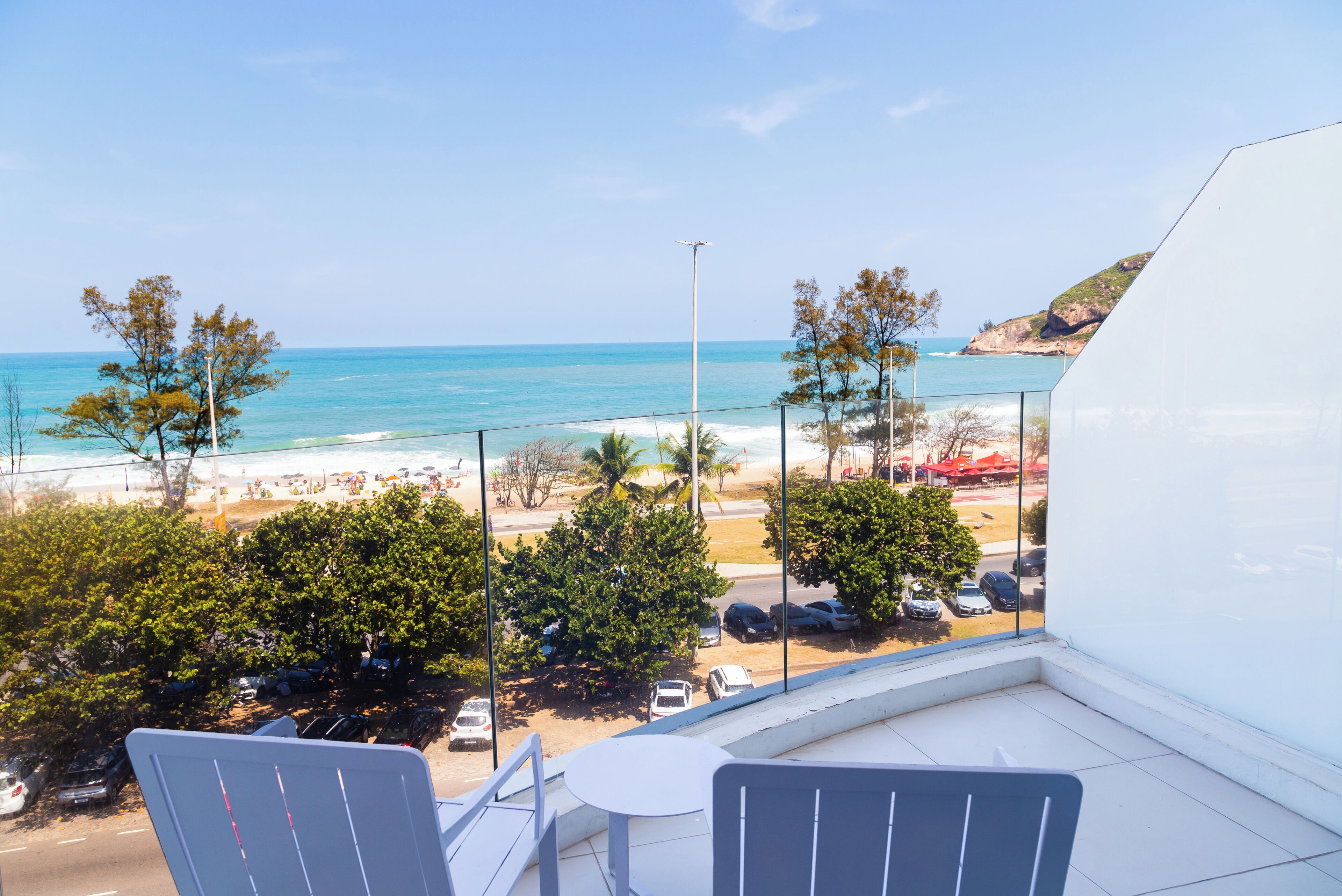 Un balcón con vista a una playa, coches estacionados y árboles.