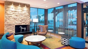 A modern lounge area with a stone wall, a fireplace, a blue sofa, a round table, and a patterned ottoman. - Fairfield Inn & Suites by Marriott Houston Pasadena (Pasadena)