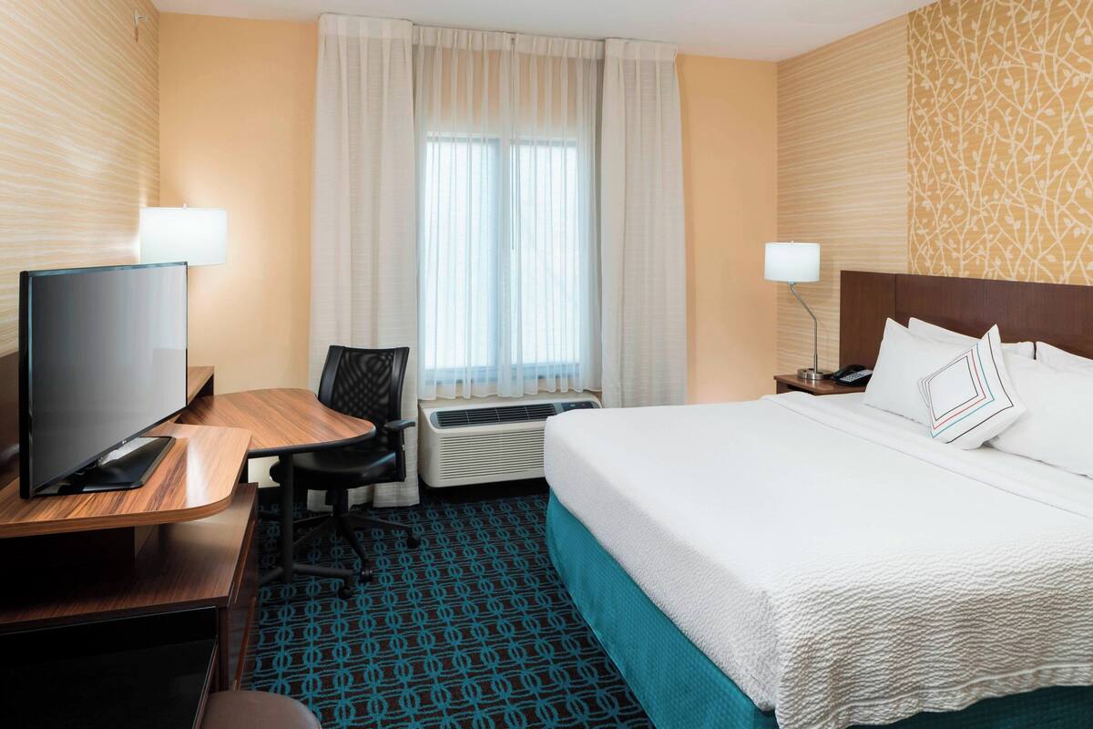 A hotel room with a bed, a desk with a TV, a chair, a window with curtains, and a lamp.