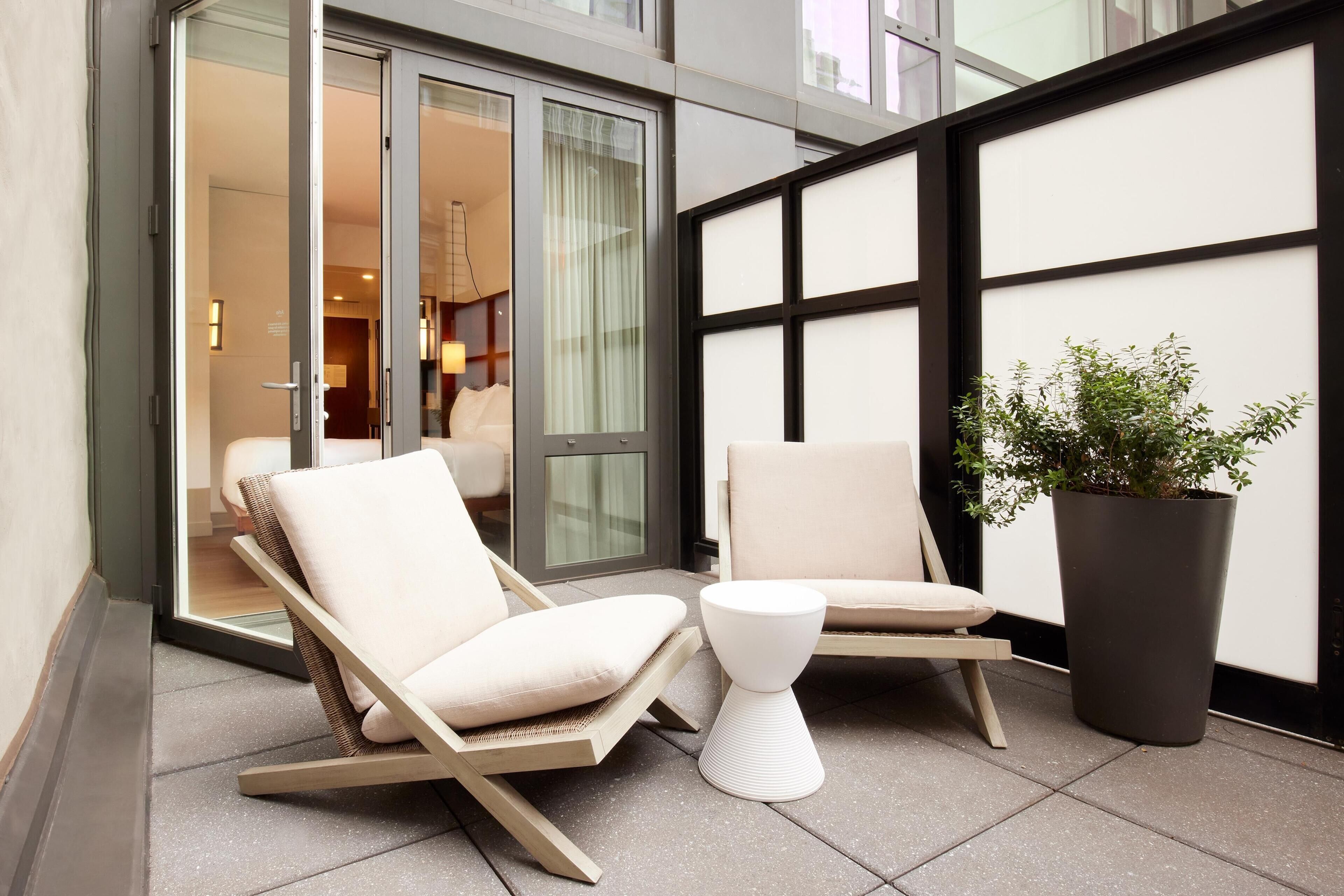 Un espacio moderno al aire libre con dos sillones reclinables, una mesita blanca, una planta en maceta y un ventanal de vidrio que da a una habitación.