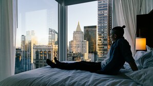 Eine Person liegt in einem Hotelzimmer auf einem Bett, durch große Fenster ist ein Stadtblick zu sehen.