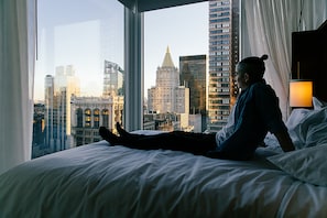 A person lying on a bed in a hotel room with a cityscape view through large windows.