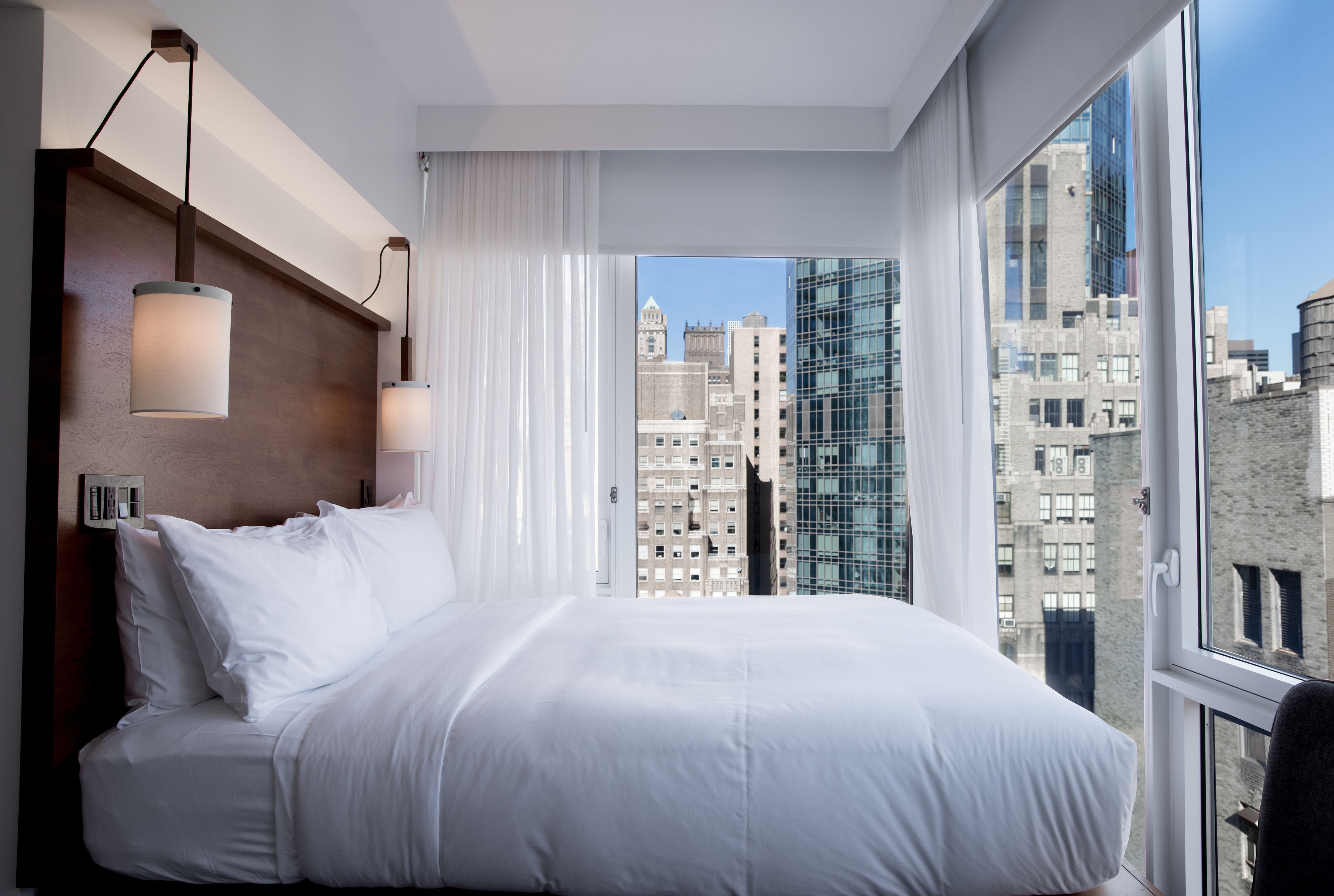 Habitación de hotel moderna con una cama grande, cabecero de madera y vistas a la ciudad a través de amplios ventanales.