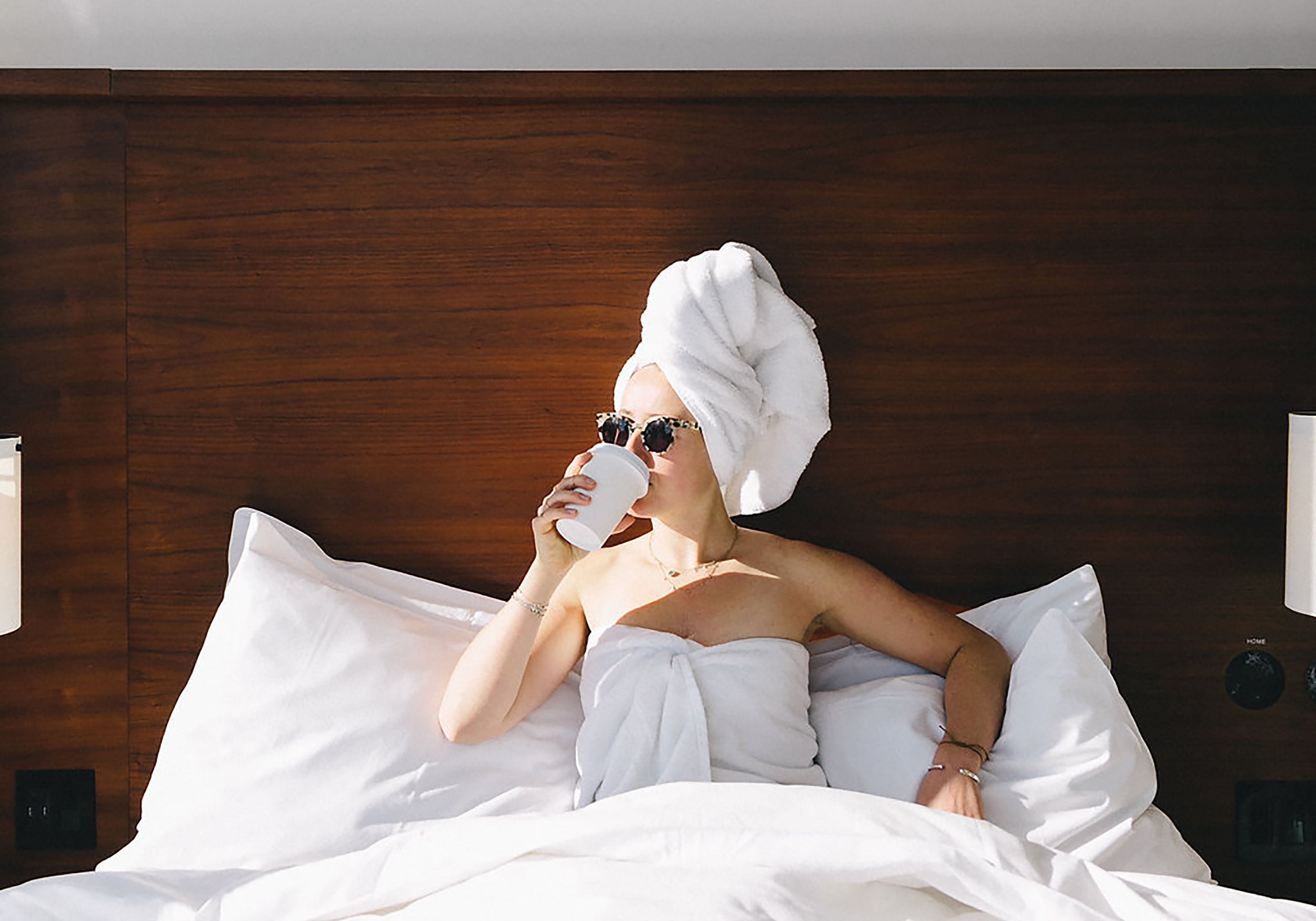 Una persona acostada en la cama con una toalla en la cabeza, sosteniendo una taza.