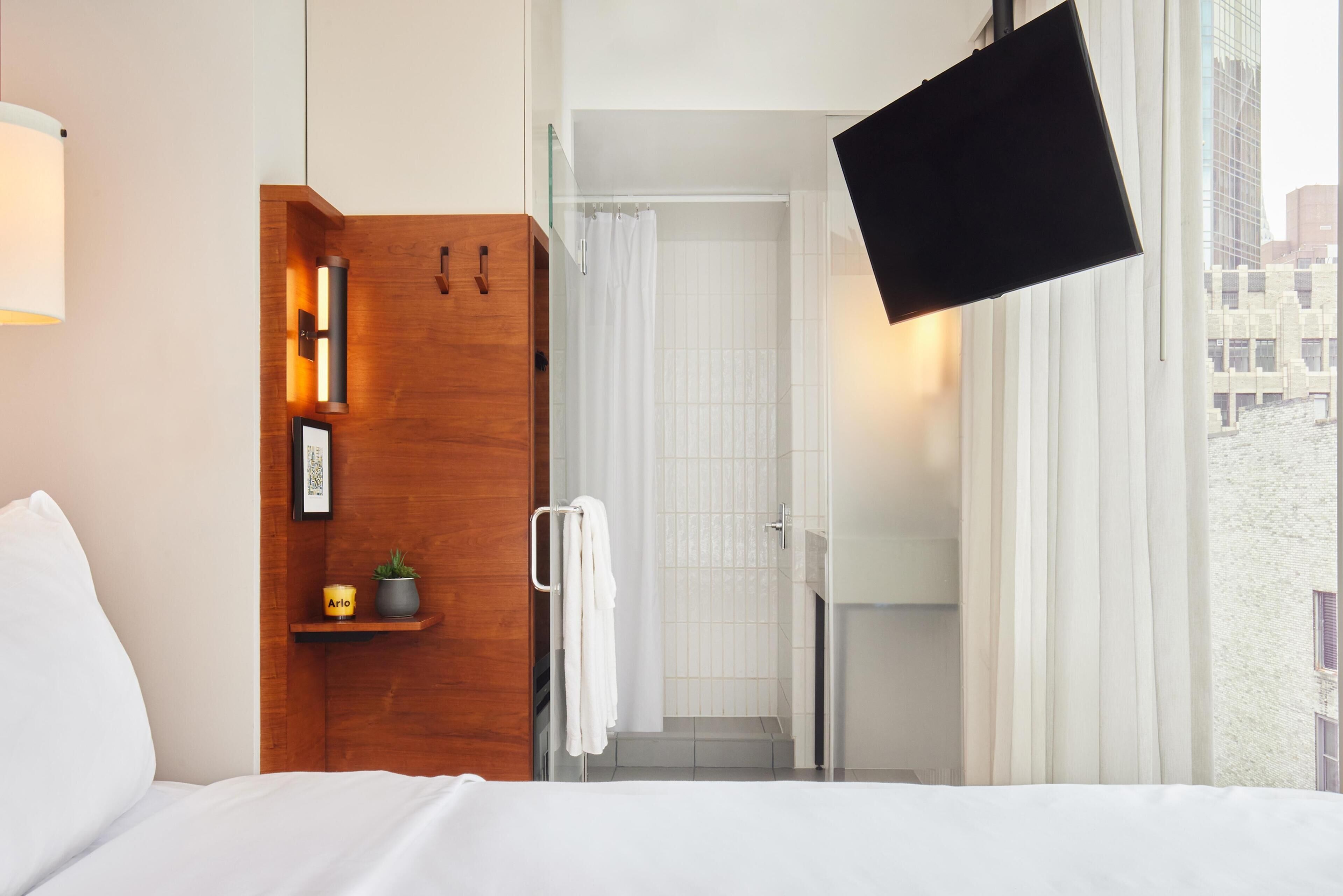 Una habitación de hotel con cama, un armario de madera con repisa, una lámpara de pared, zona de ducha y ventana con cortinas.