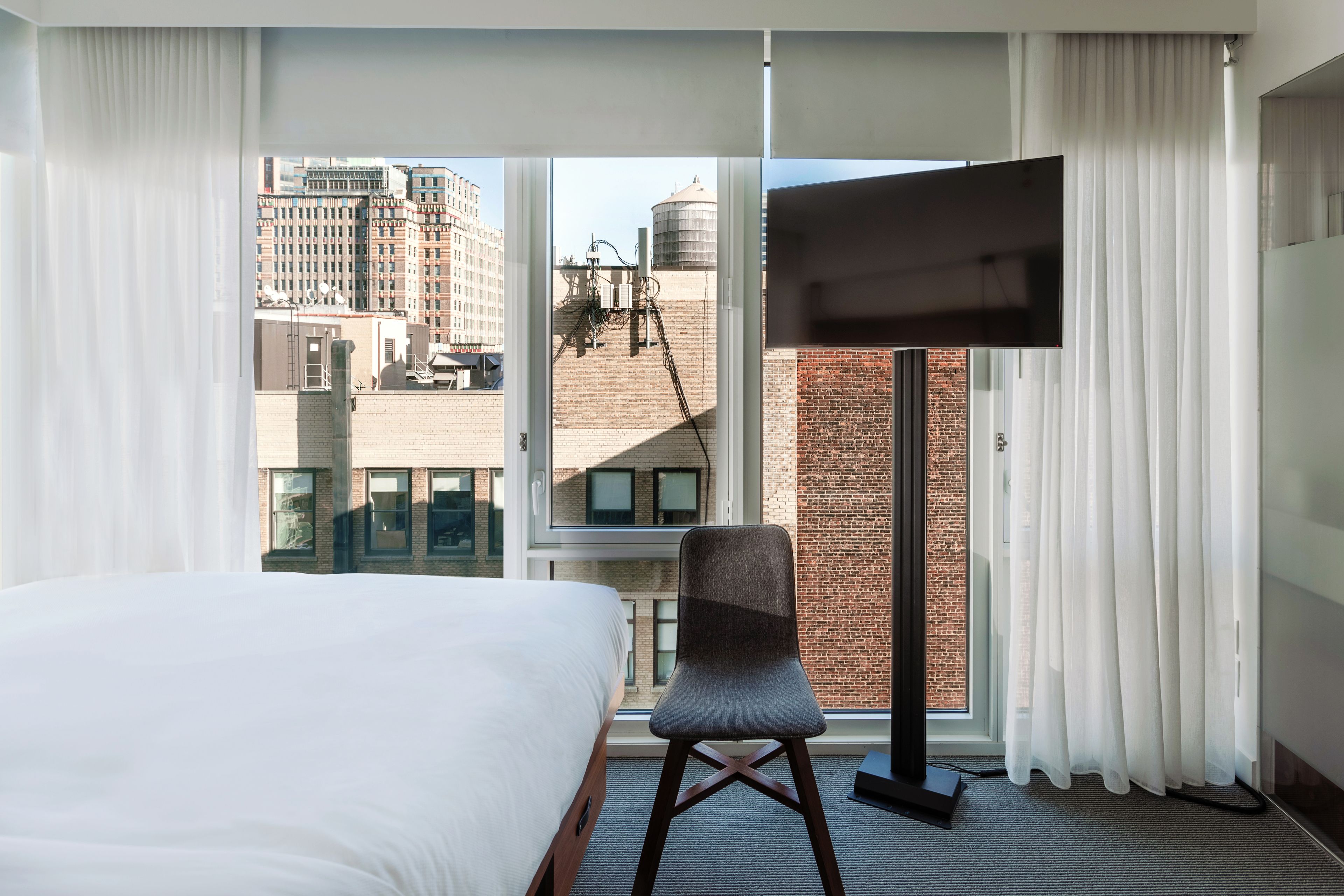 A hotel room with a large bed, a chair, a flat-screen TV, and a view of the cityscape through a large window.